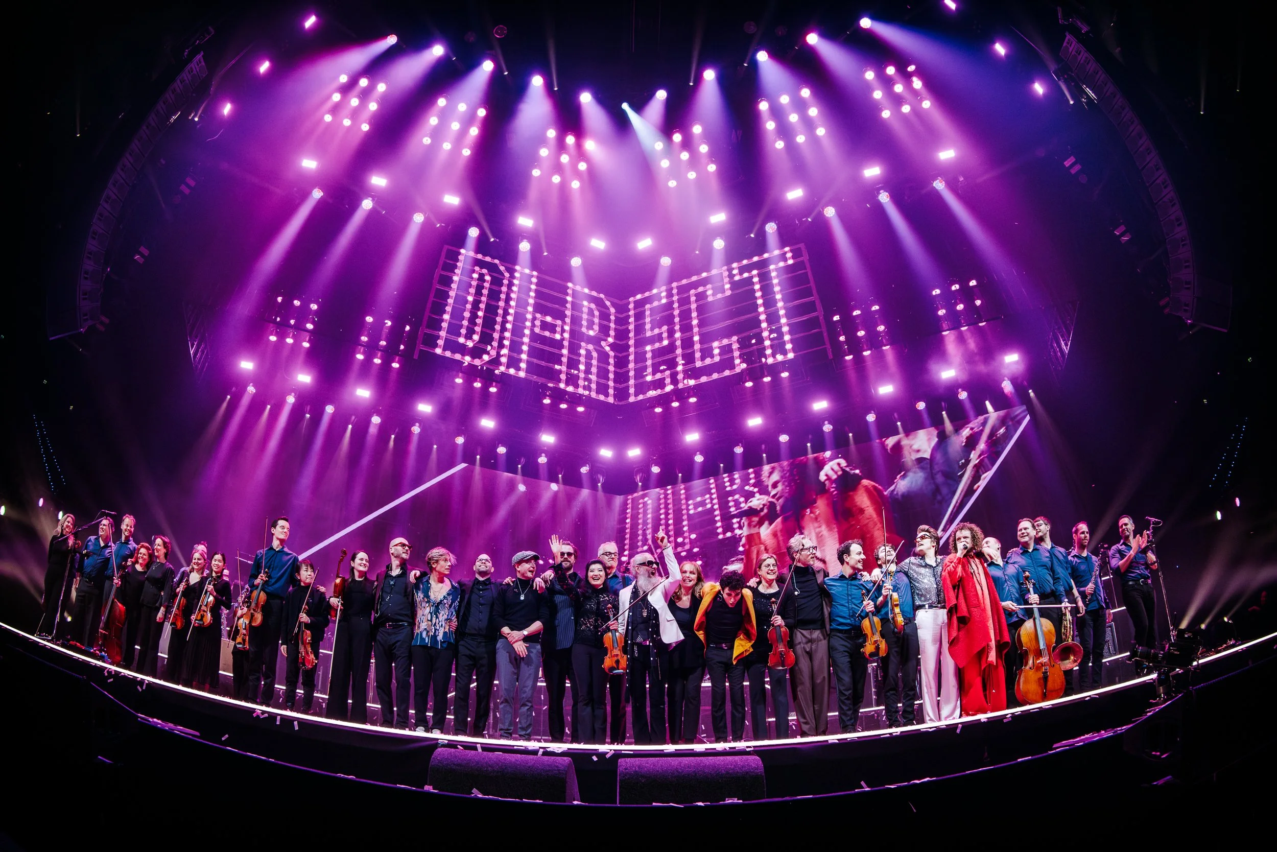 Large orchestra on stage during a concert, with purple stage lighting and a digital sign showing the word 'DI-RECT' overhead.