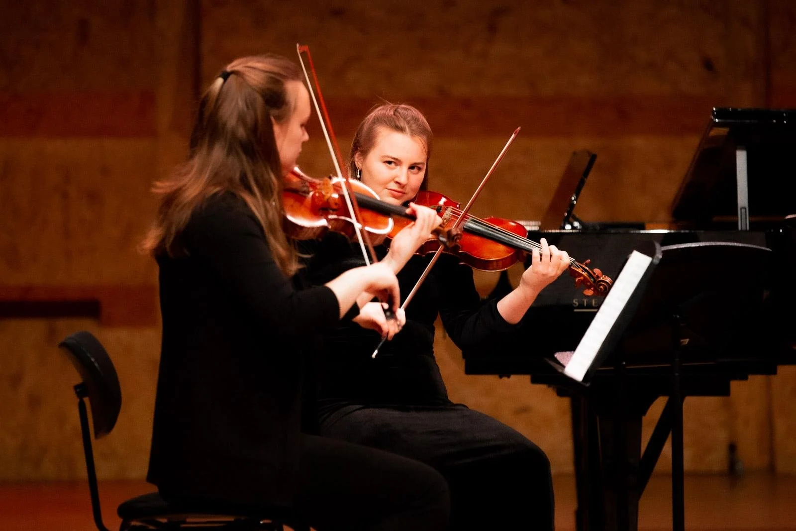 Two young women playing violins during a musical performance on stage with a grand piano in the background.