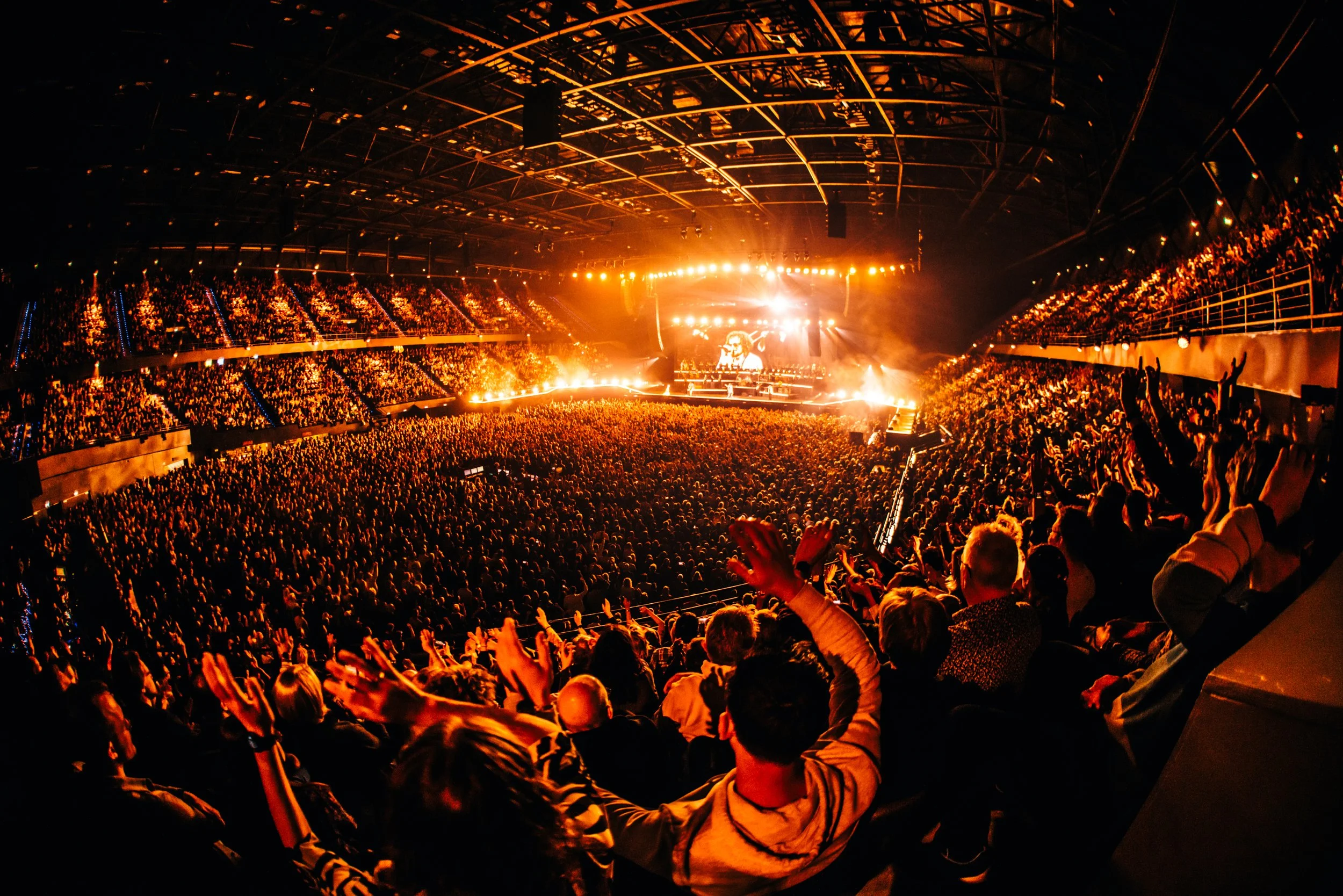 A large indoor concert or music festival with a packed crowd, bright stage lights, and a stage with performers, under a high roof structure.