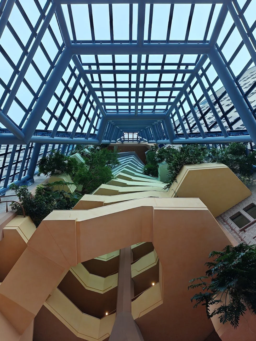 Binnenhof van een modern gebouw met meerdere verdiepingen, gekleurde muren, groene planten, en een glazen plafond.