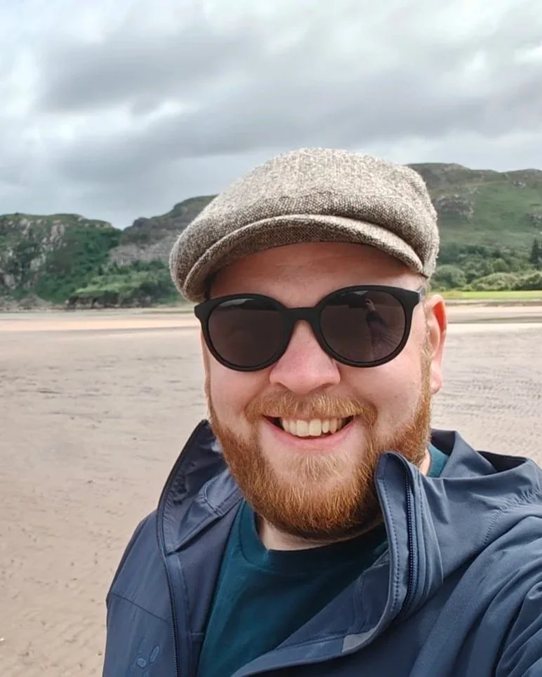Man met rode baard, zwarte zonnebril, grijze hoed en blauwe jas op strand met heuvels en bewolkte hemel op achtergrond.
