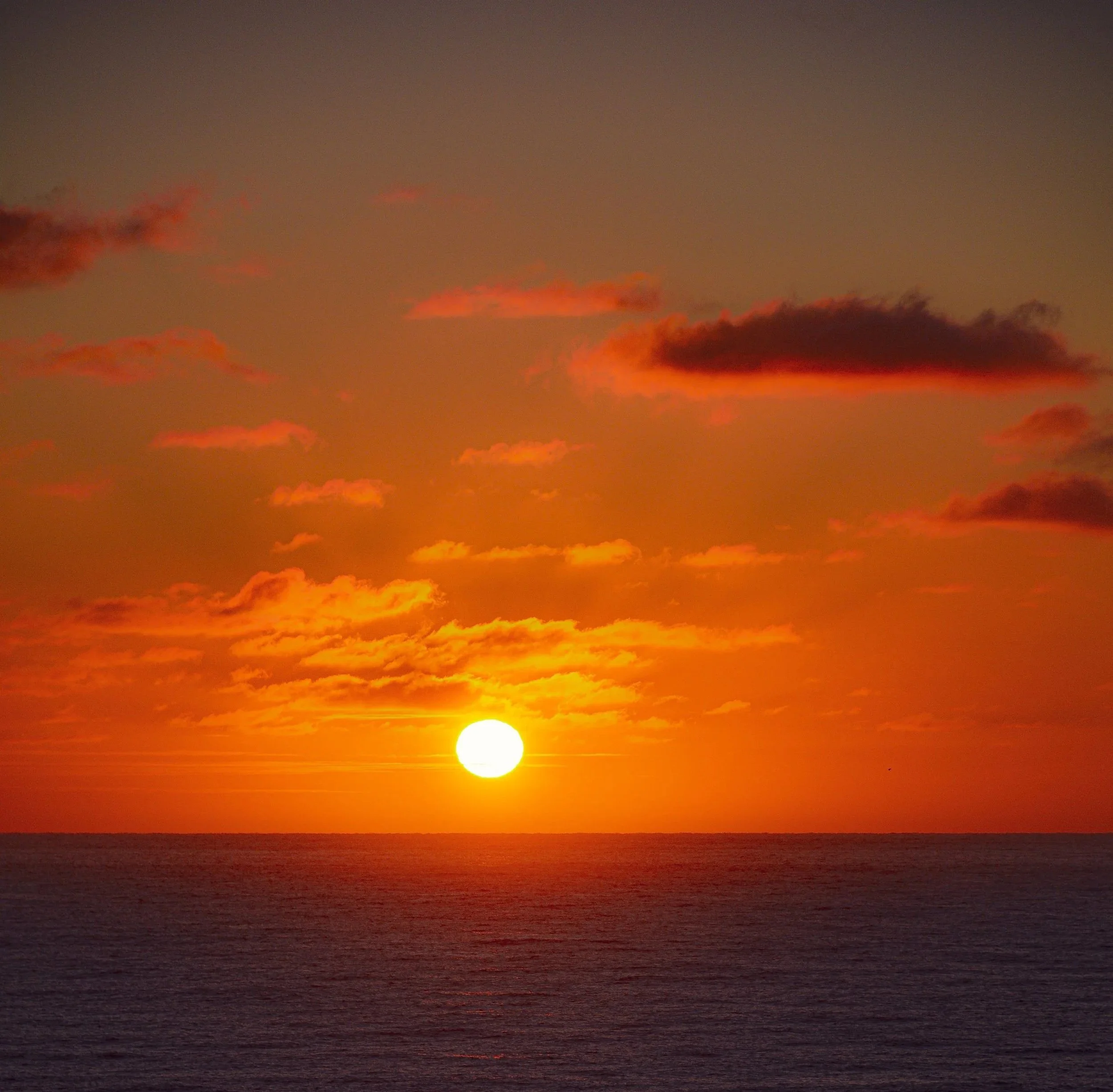 Sun setting over the ocean with a vibrant orange and red sky and scattered clouds.