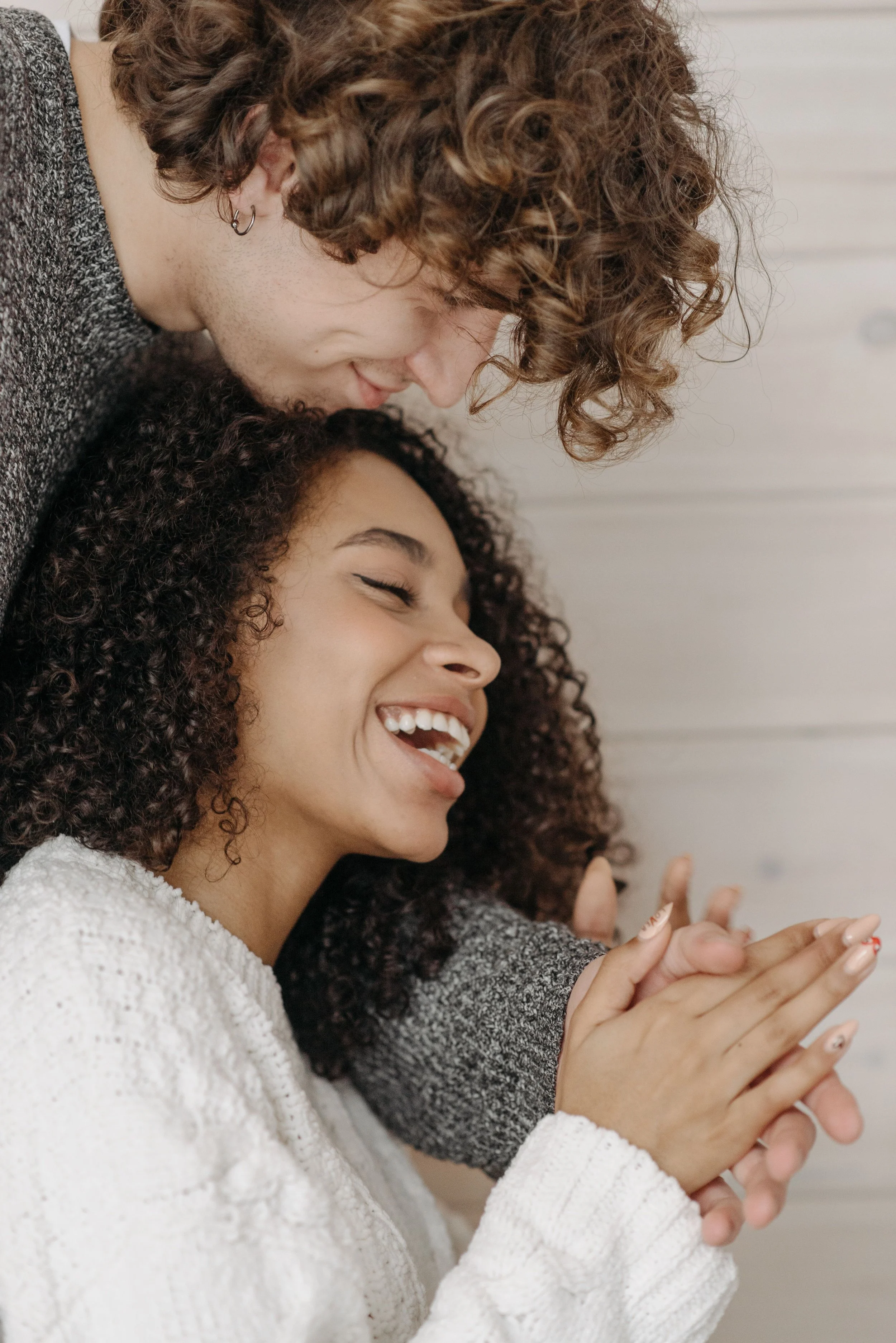 Two women with curly hair sharing a joyful moment, smiling and leaning close together. One woman has dark brown hair, the other has reddish-brown hair. They are dressed casually, with one wearing a white textured sweater and the other in a gray sweater. They are indoors against a light-colored background.
