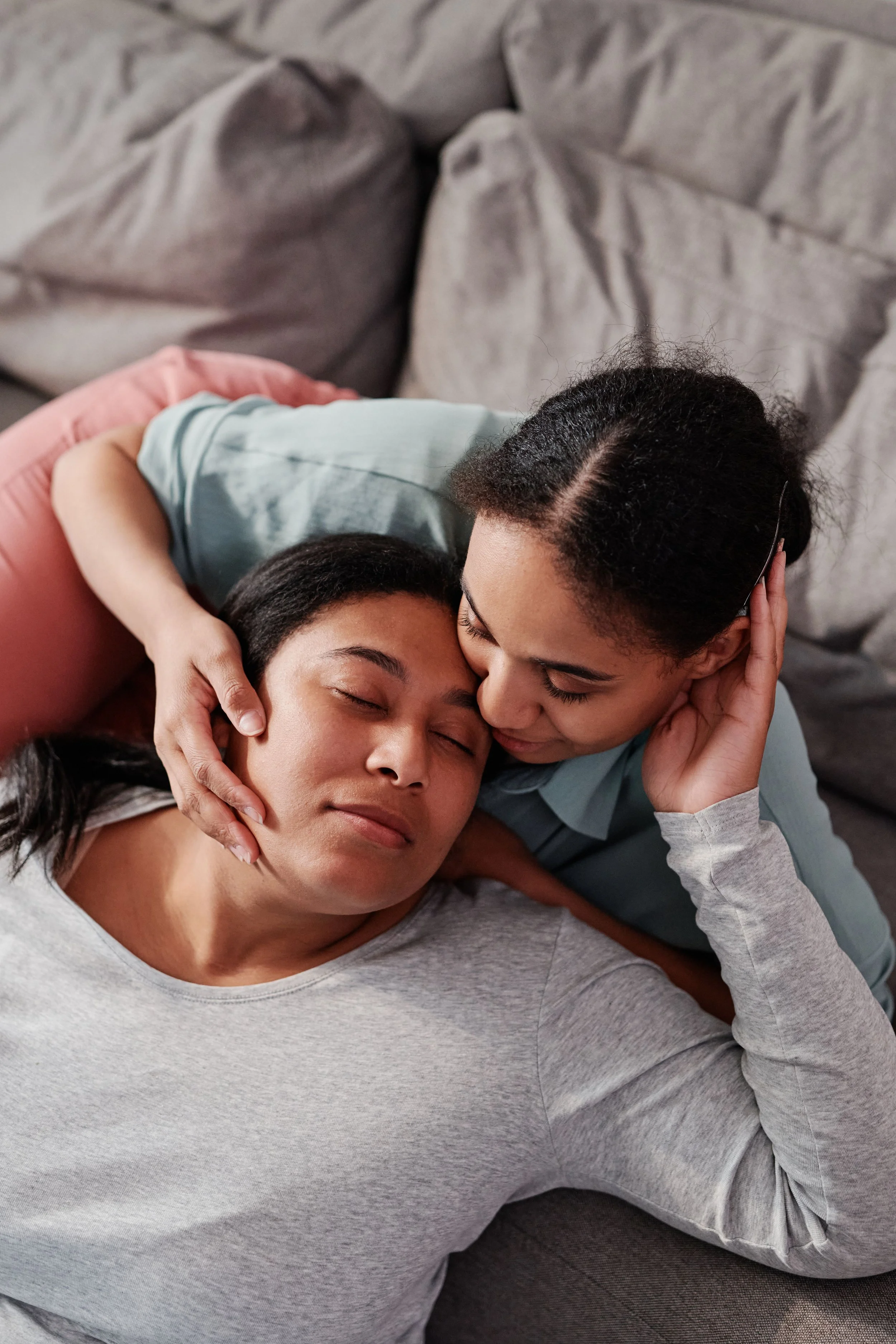 Two women sharing a tender moment, lying down together with their eyes closed, one gently holding the other's head.