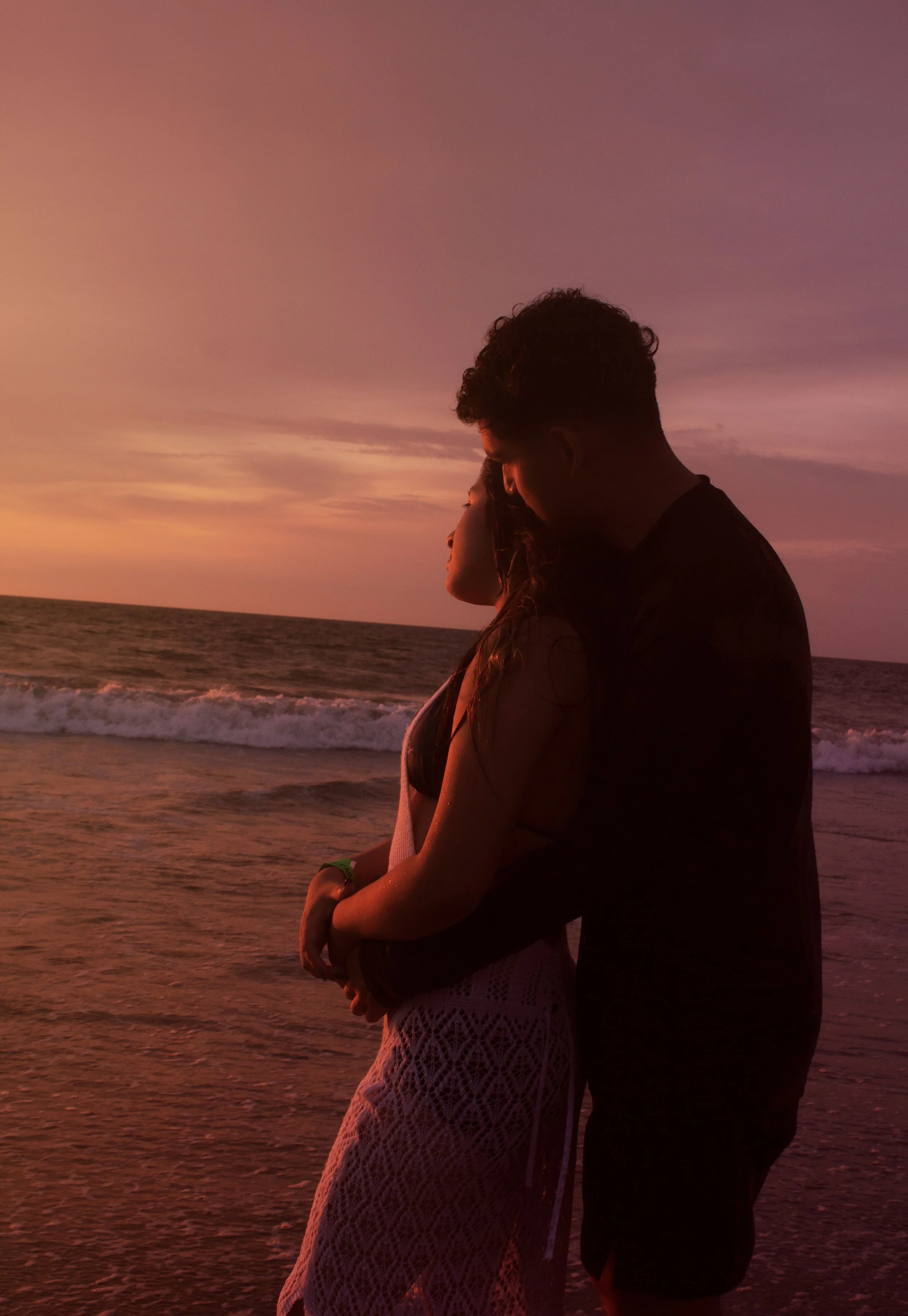 A couple sits closely together on a beach during sunset, with ocean waves in the background, sharing an intimate moment.