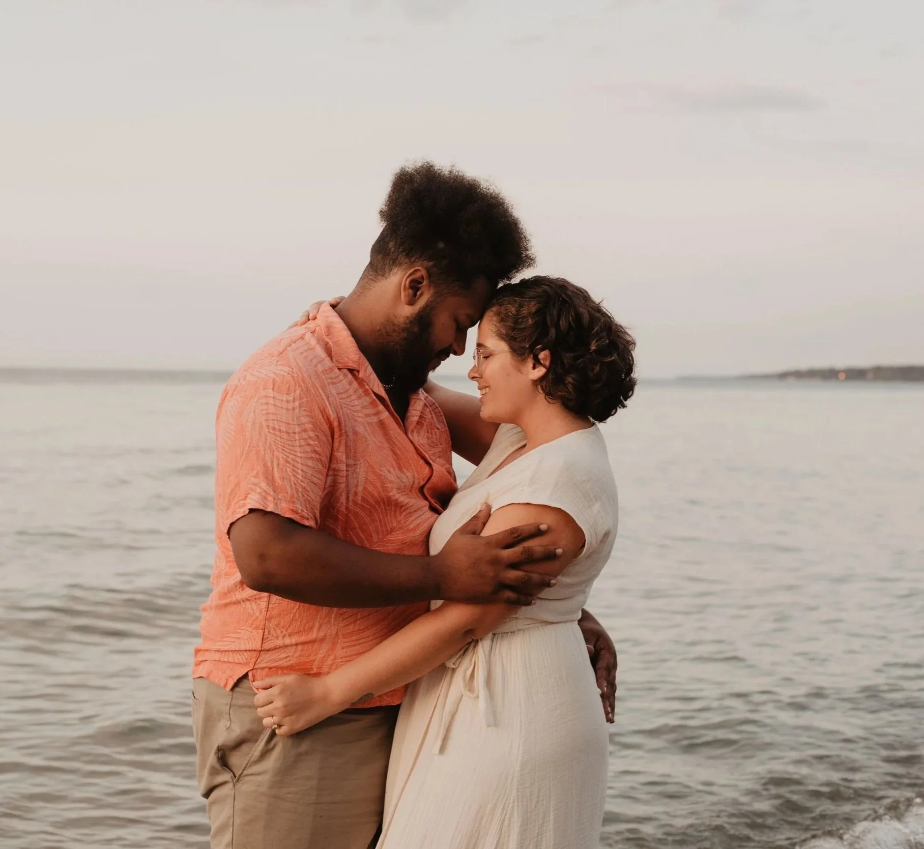 A couple embraces on the beach, with the ocean and a cloudy sky in the background. The man is wearing a salmon shirt and beige shorts, and the woman is wearing a white dress and glasses. They are touching foreheads and smiling.