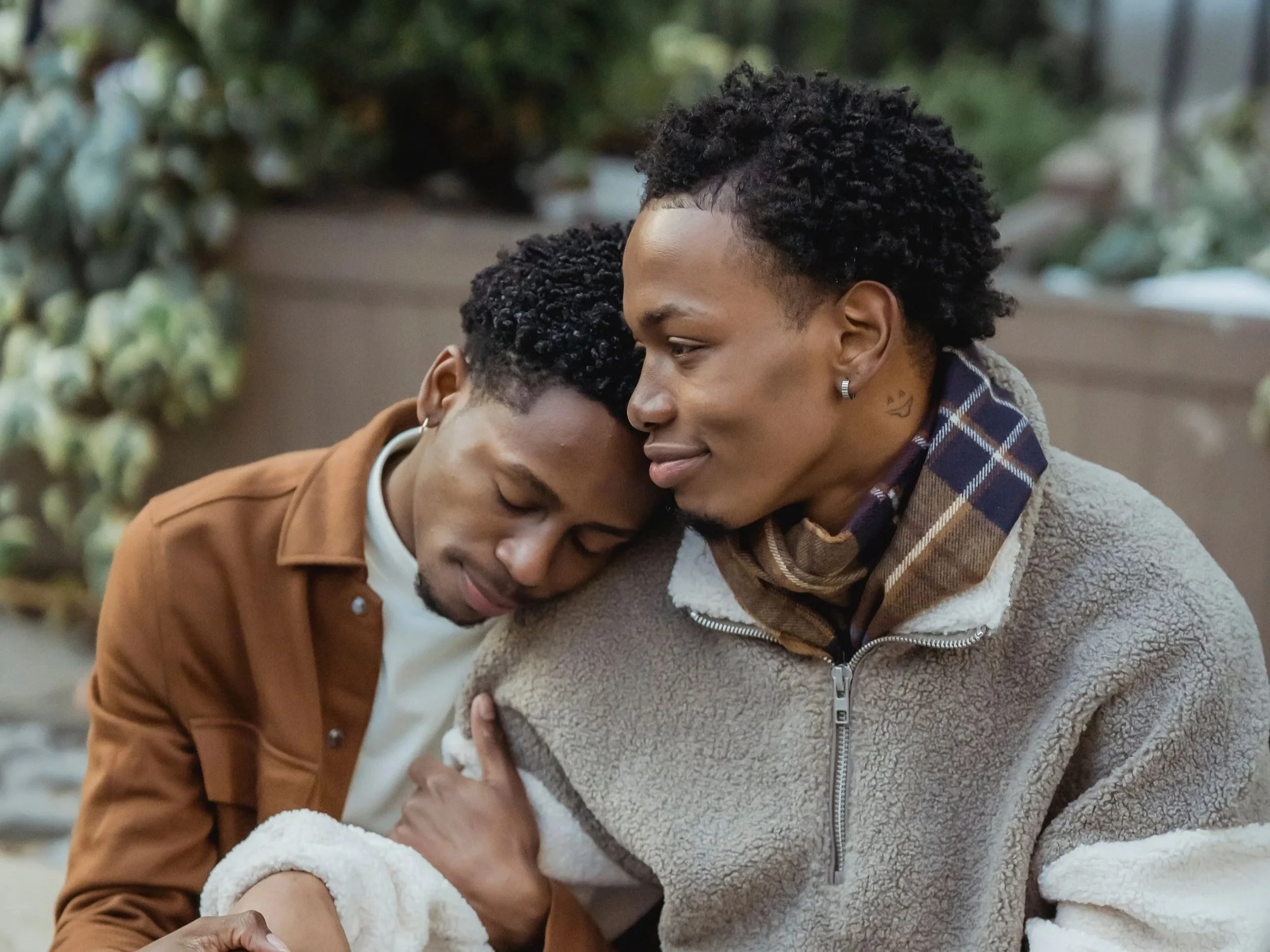 Two people sharing an intimate moment, with one resting their head on the other's shoulder in a park or outdoor setting.
