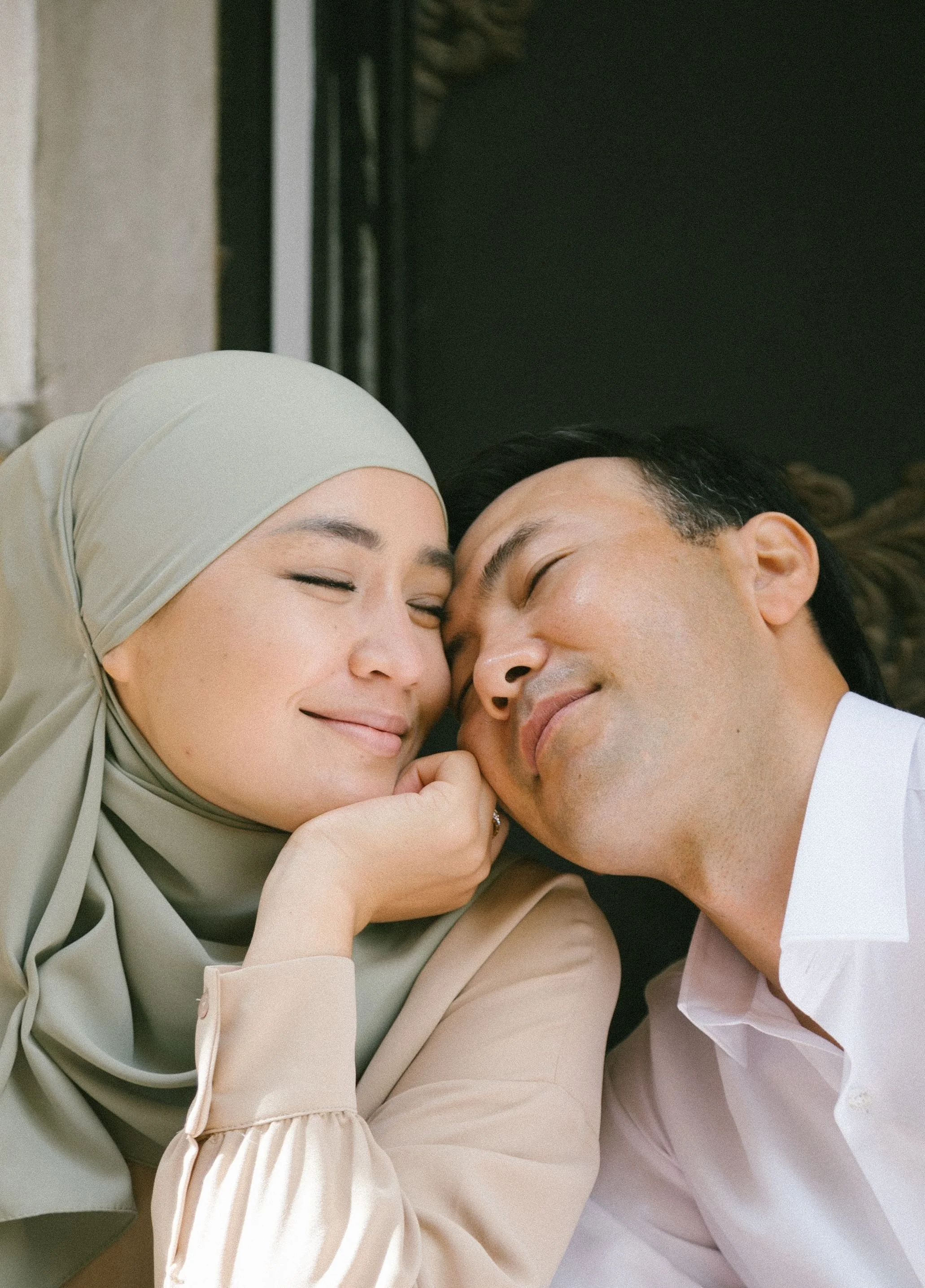 A woman wearing a beige hijab and a beige long sleeve top is lying on a bed with a man with short black hair and a light pink shirt, both with eyes closed and smiling.