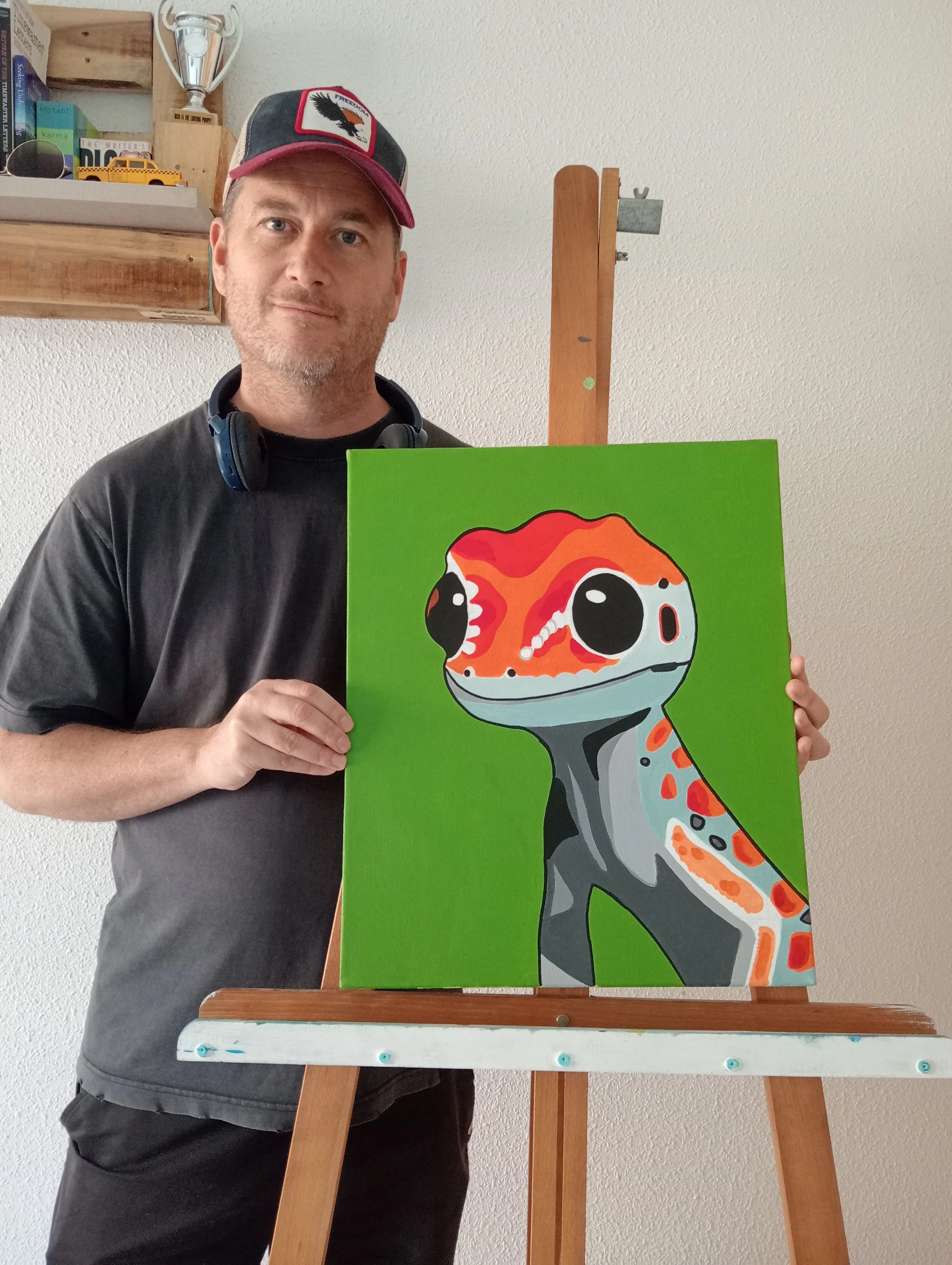 A man wearing a red and black cap with a logo of a bird and the word "FREEDOM" on it, holding a painting of a colorful, stylized gecko on a bright green background, standing in front of an easel.