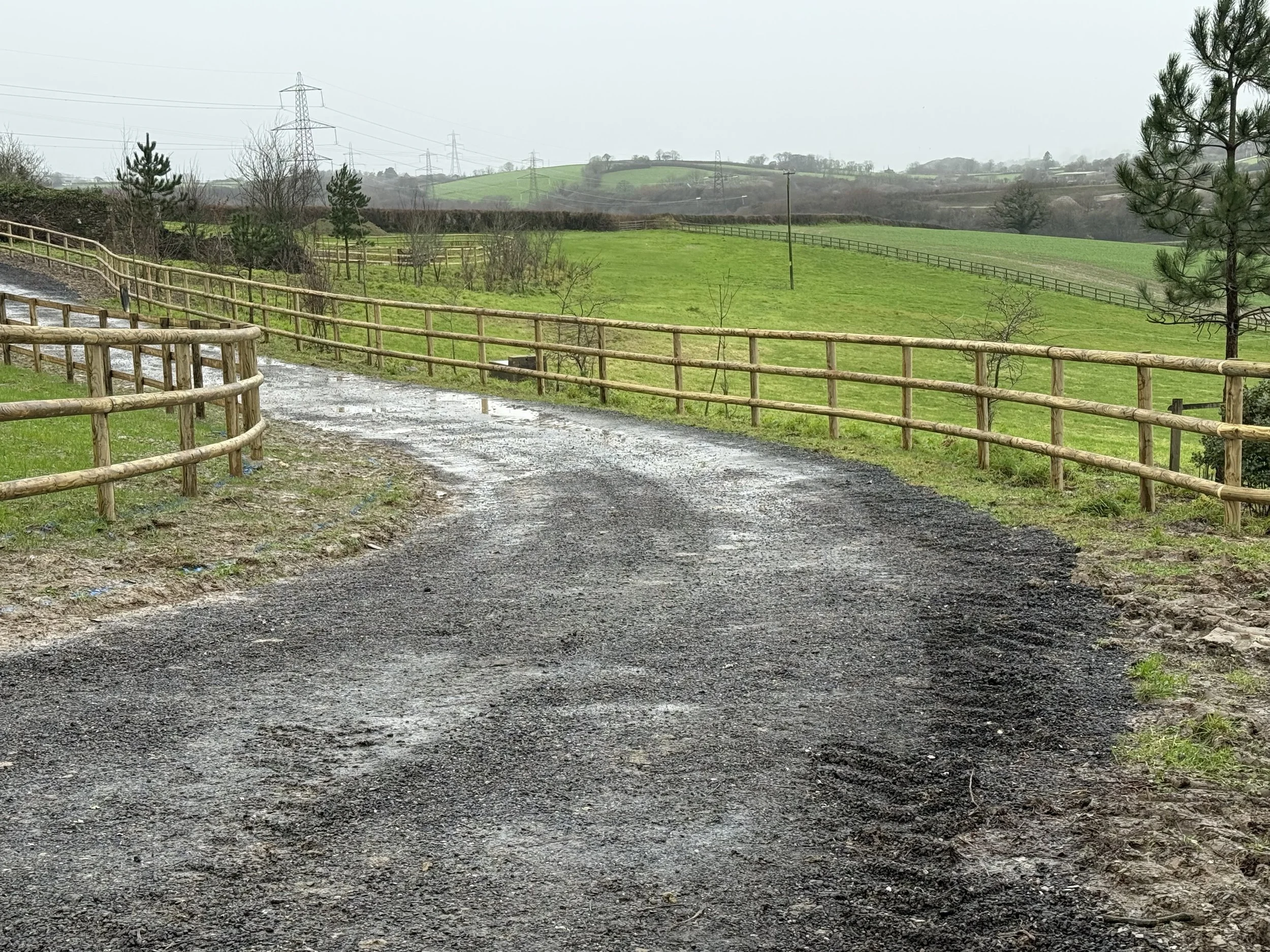 A dirt and gravel pathway bordered by wooden fences, leading into a lush green field with scattered trees and rolling hills in the distance, overcast sky.