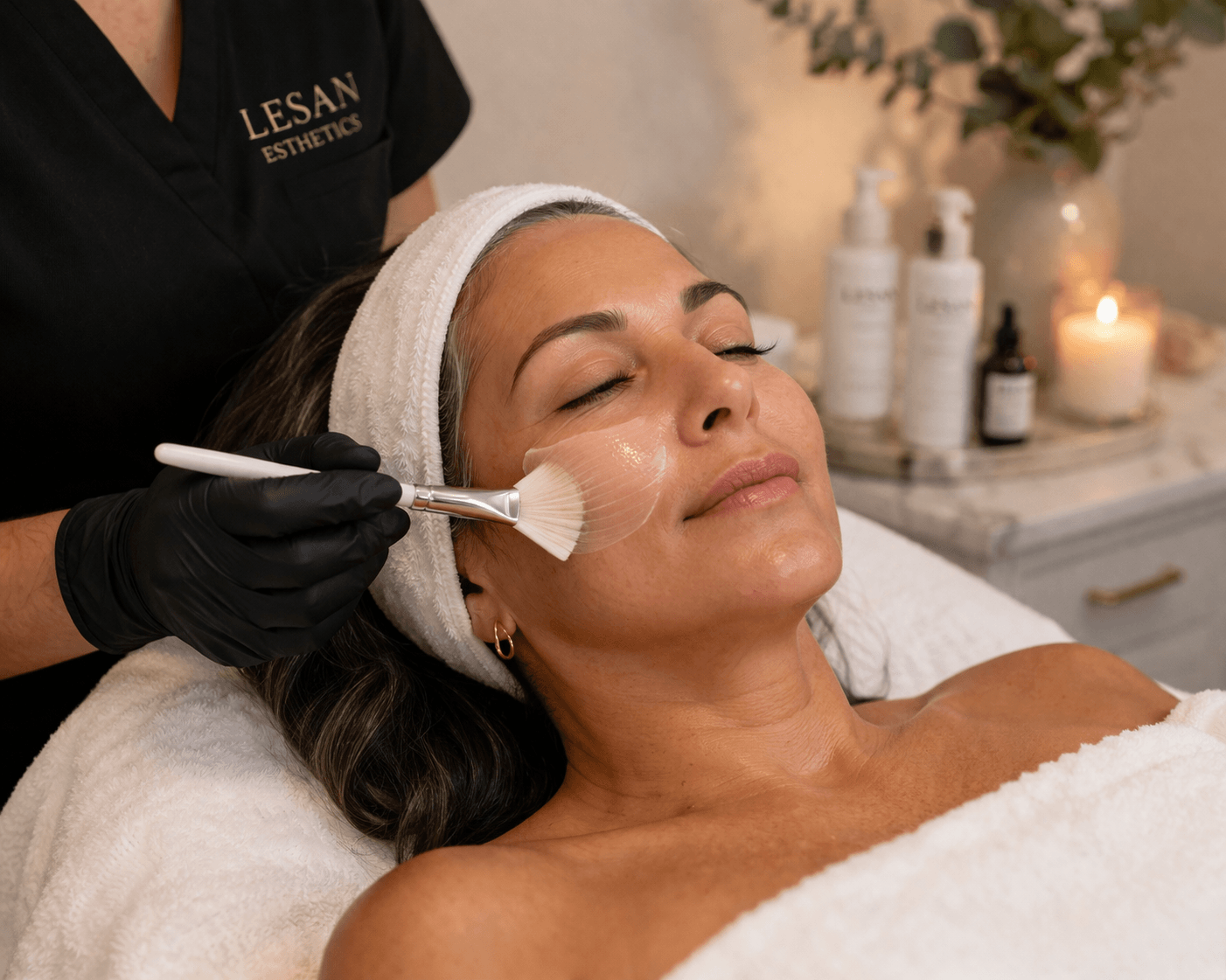 A woman receiving a facial treatment at a spa, lying down with her eyes closed, a towel wrapped around her head, and a skincare product being applied with a brush. The background shows skincare bottles and a lit candle.