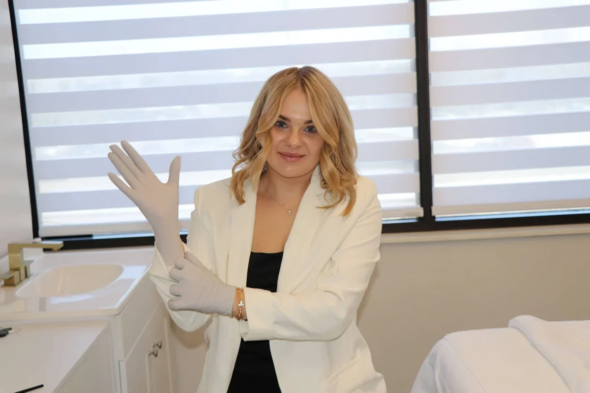 A woman with blonde hair in a white blazer and black top putting on medical gloves in a clinical setting with windows and blinds in the background.