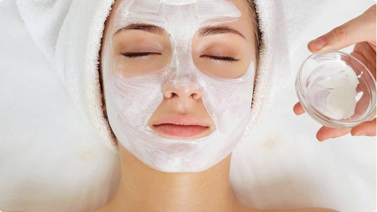 A woman with a towel wrapped around her head is receiving a facial treatment, with her eyes closed. A white cream mask is being applied to her face, and a hand is holding a small bowl of the white cream.