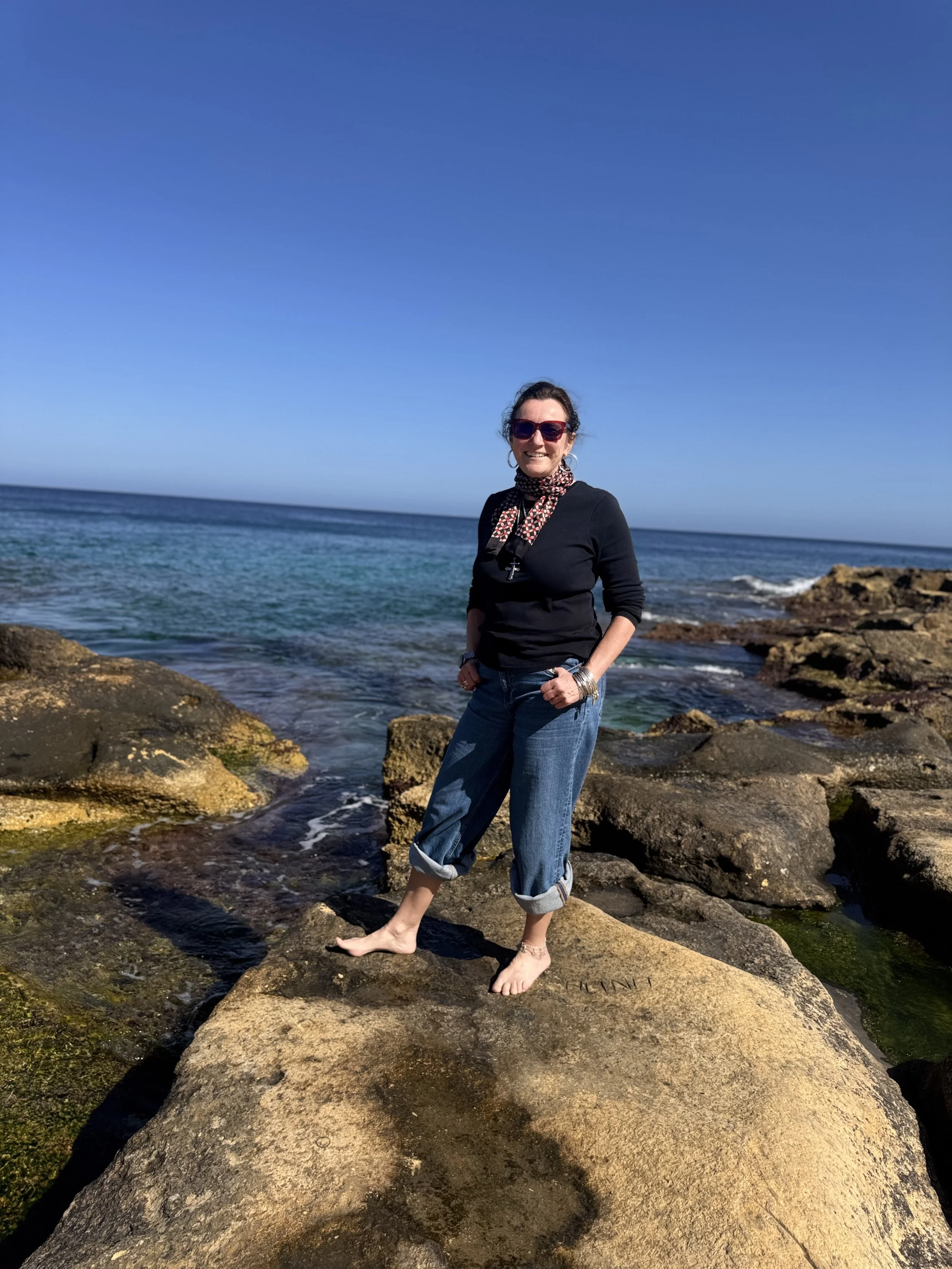A woman with dark hair and sunglasses standing barefoot on rocks by the ocean, wearing a black long-sleeve T-shirt, jeans with rolled-up cuffs on the legs, and a patterned scarf, smiling at the camera against a bright blue sky.