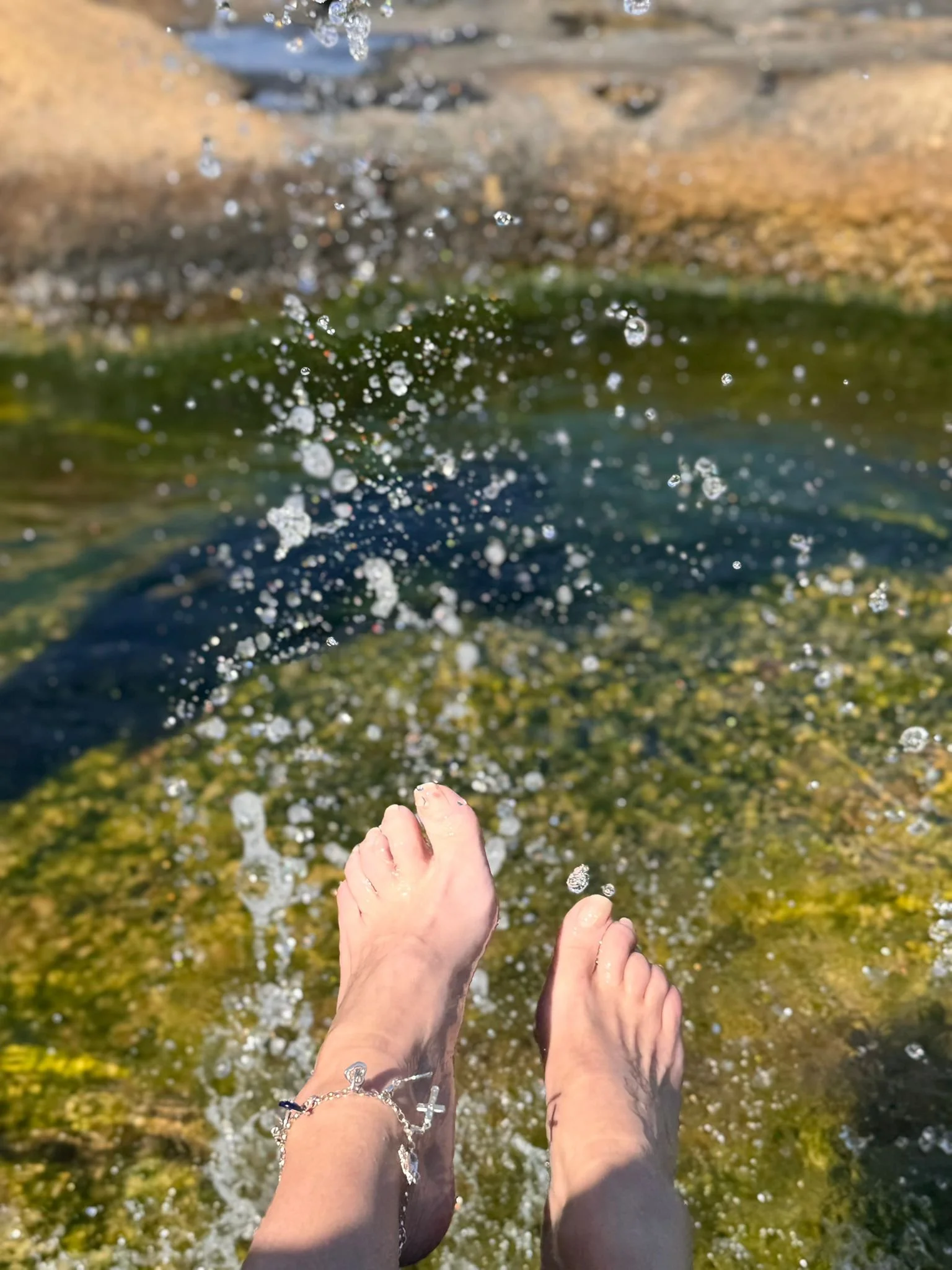 A woman bathing feet in clear water with rocks and greenery in background, jewellery on ankle.