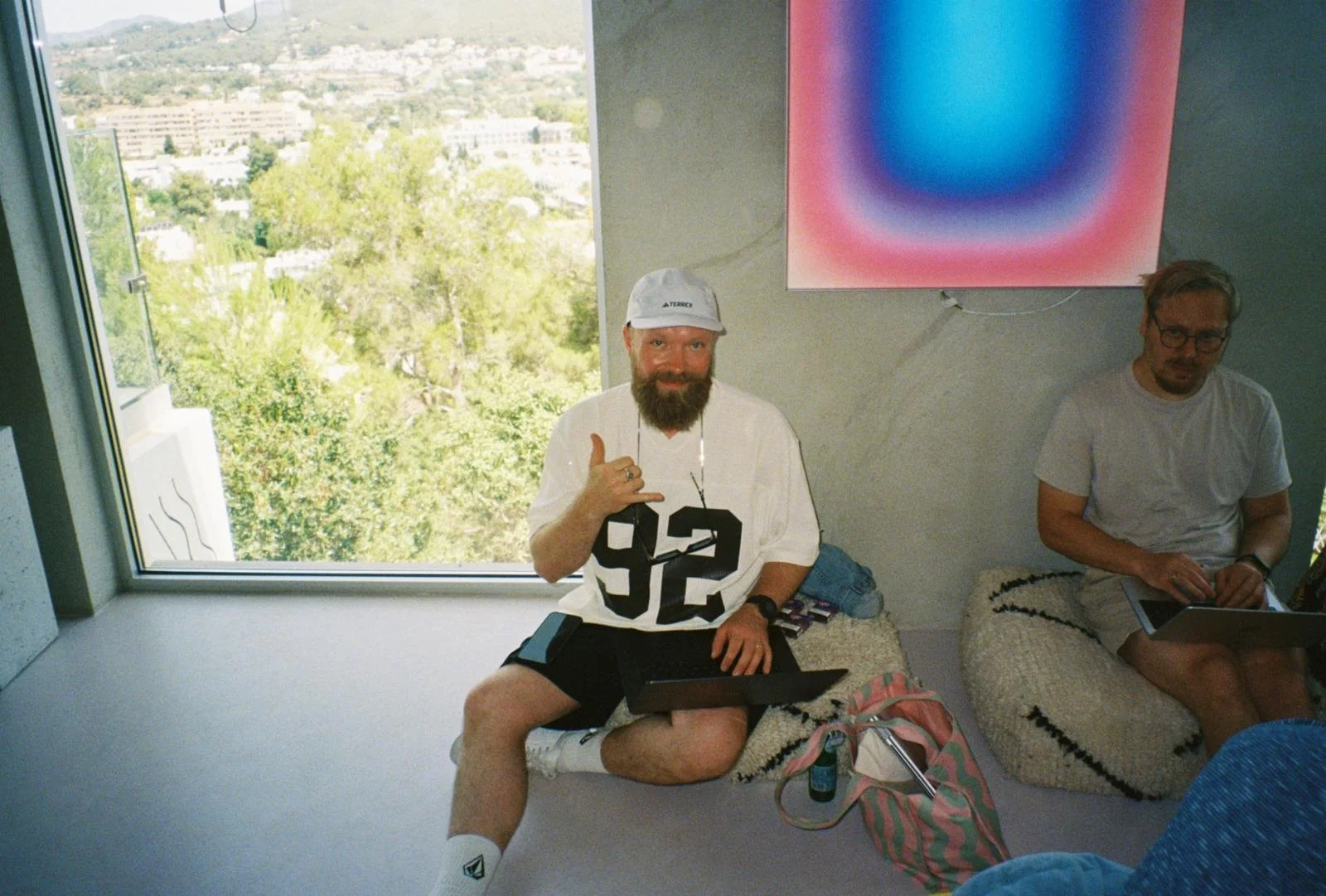 Two men sitting indoors near a large window with a view of trees and buildings outside. One man has a beard, is wearing a white shirt with black numbers, shorts, a white cap, and is making a hand gesture while smiling. The other man has glasses, a beard, and is wearing a white t-shirt, looking at a tablet. There are bags, a bottle, and electronic devices on the seats they are sitting on.