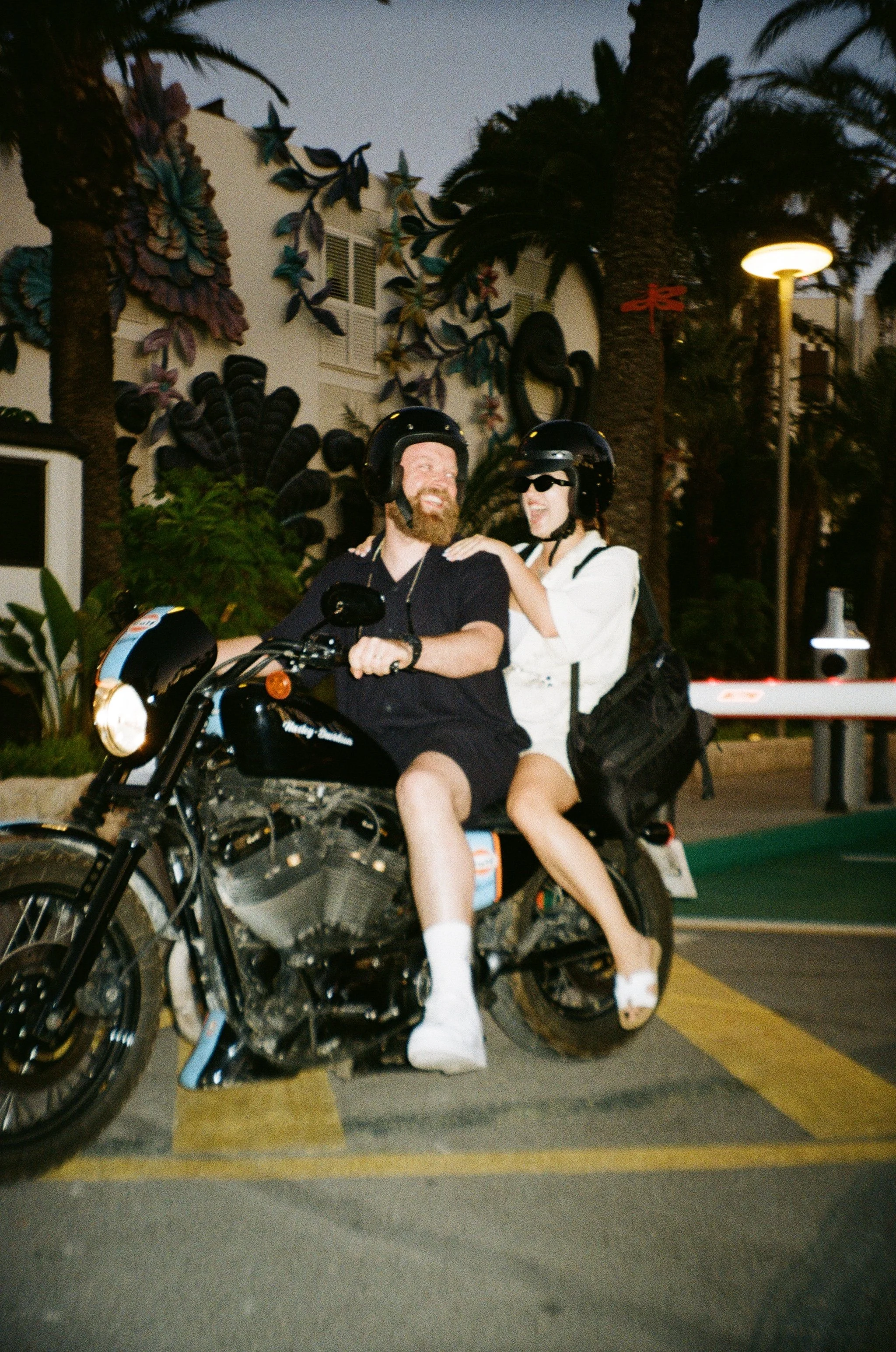 Two people riding a motorcycle in a parking lot with palm trees and decorative wall art in the background at dusk. The rider wears a black helmet and black shirt, while the passenger wears a black helmet, white shirt, sunglasses, and is barefoot.
