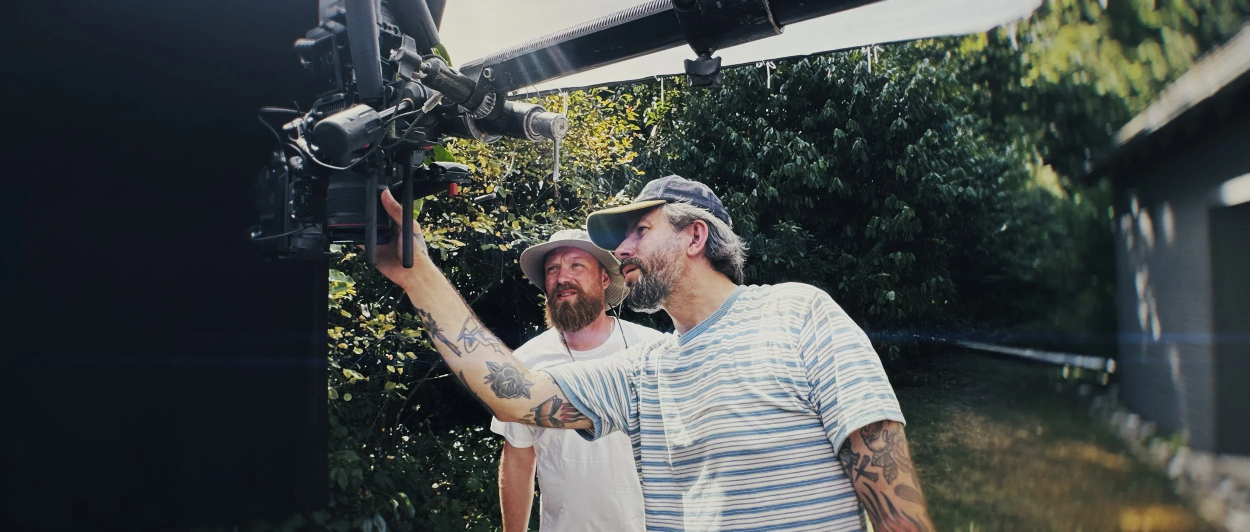 Two men working on a film camera outdoors amid trees, one adjusting the camera, the other observing, both wearing hats and casual clothing.