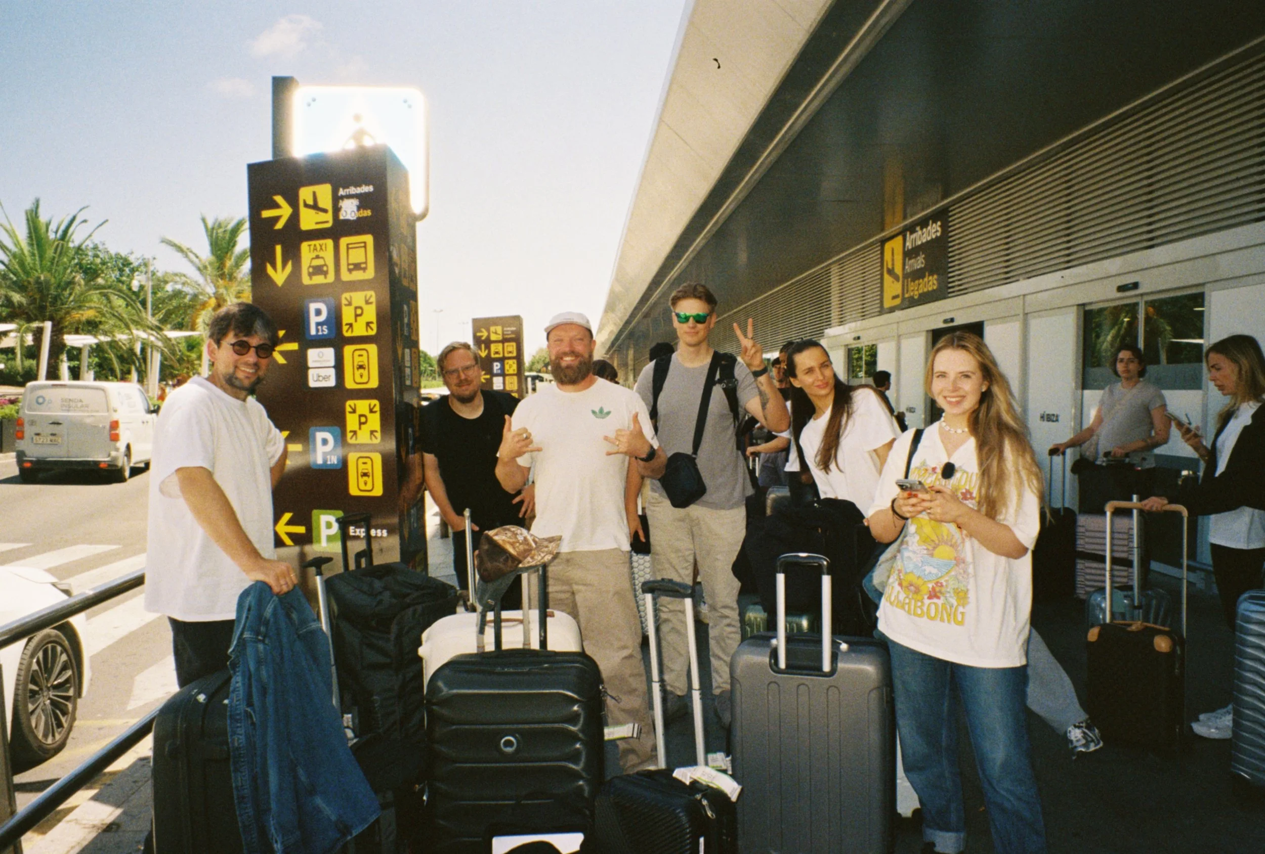 A group of six young adults smiling and posing with luggage at an airport terminal, with signs indicating arrival, departure, taxis, and parking in the background.