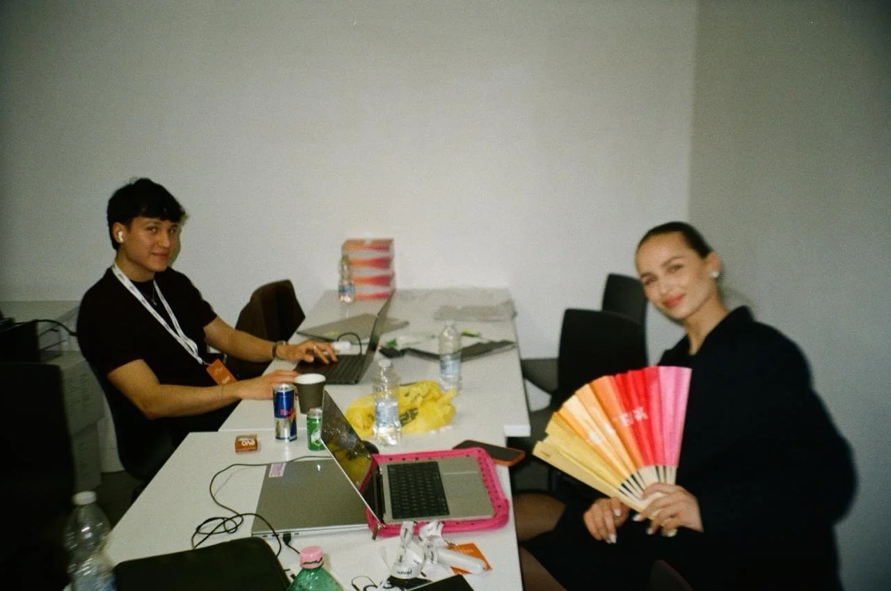 Two women working at a conference table with laptops, water bottles, and snacks; one woman is using a laptop, the other is holding a colorful folding fan.