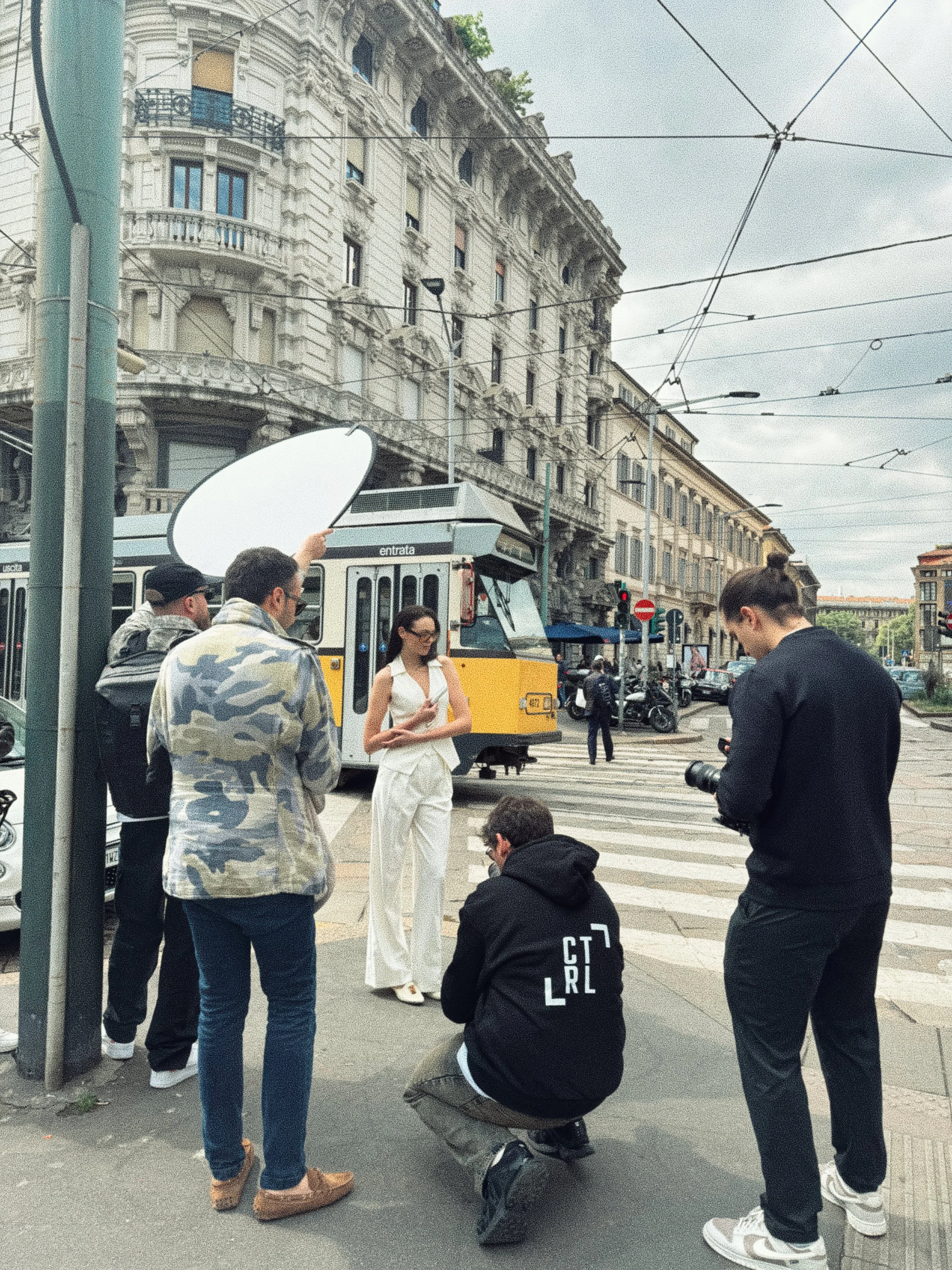 A group of people gathered on a city street corner with some standing and one kneeling, three taking photos or videos, a woman in white pants and others in casual clothing, with urban buildings, streetcar tracks, and a bus in the background.