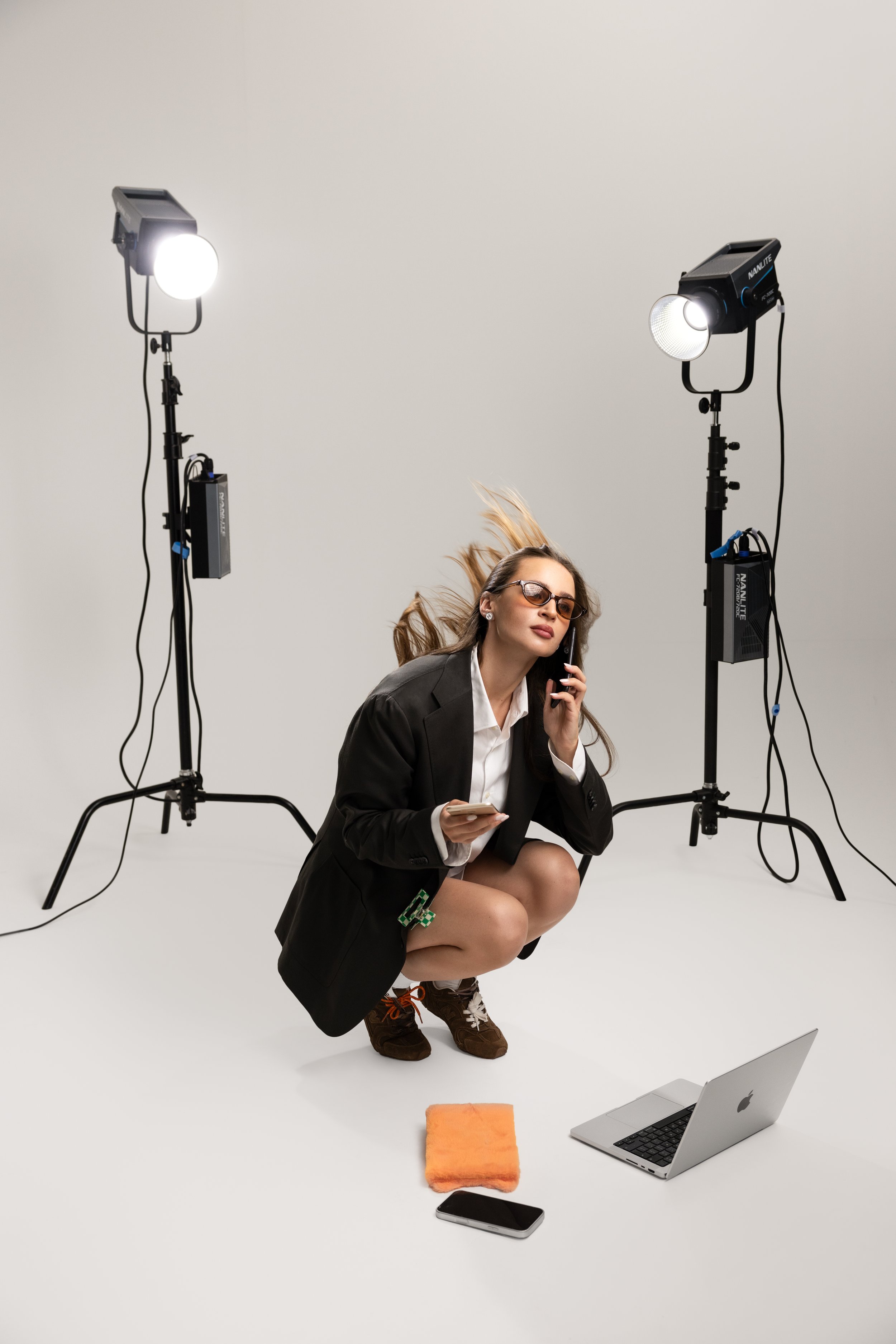 A woman in business attire squatting in front of a plain backdrop, holding a smartphone to her ear. She is surrounded by professional lighting equipment, a laptop, a smartphone, and other objects on a white surface, suggesting a photoshoot or video recording setting.