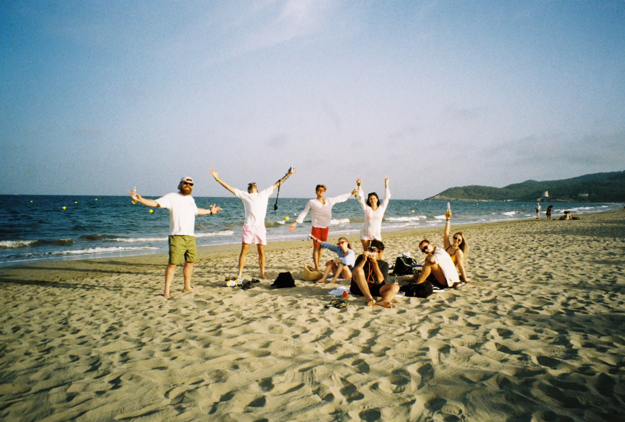 A group of people enjoying a day at the beach, standing and sitting on the sand with the ocean and distant hills in the background.