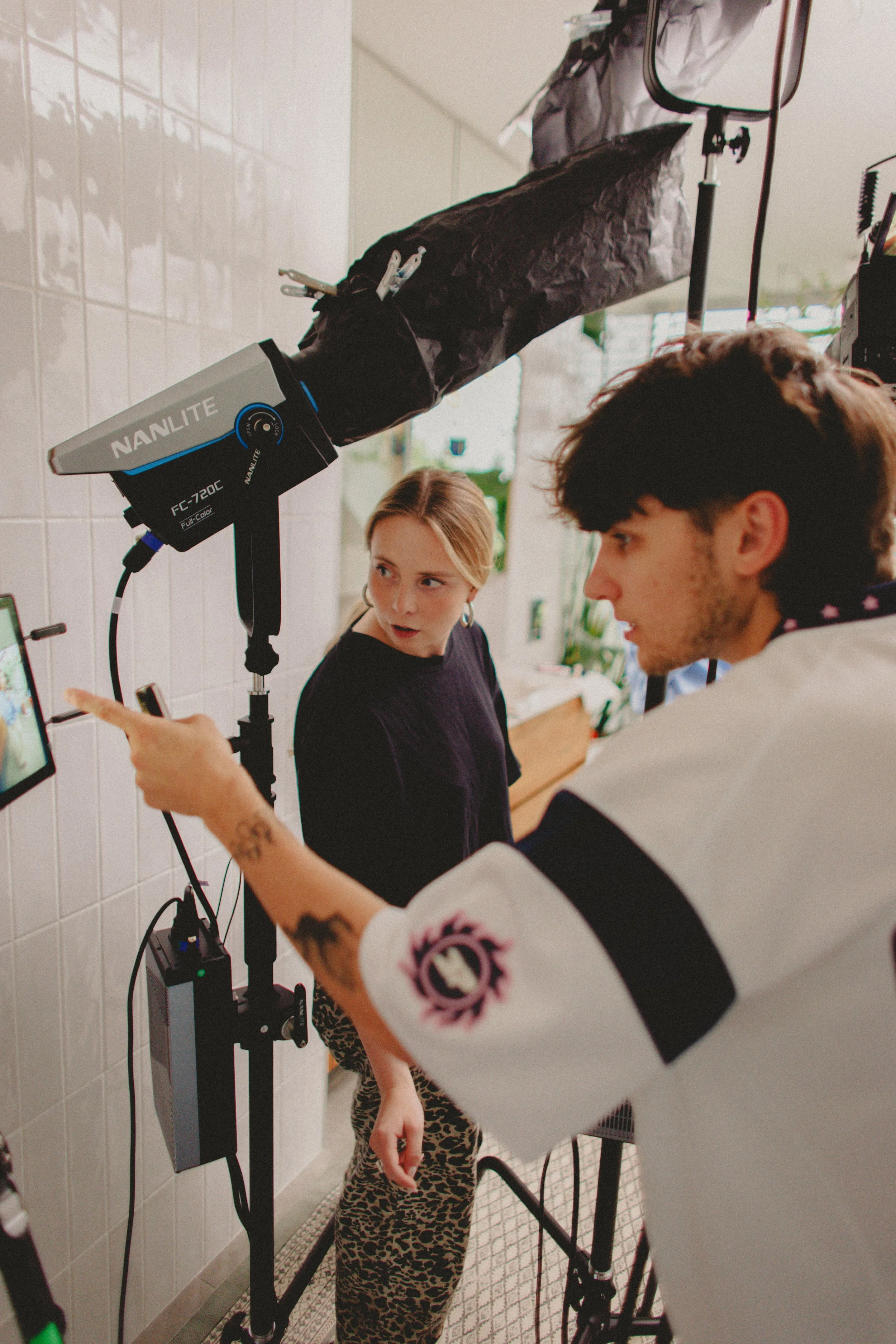 A woman and a young girl with leopard print pants look at a monitor while a professional using a scanning device scans the woman's arm for a skin analysis in a room with white tiled walls and natural light.