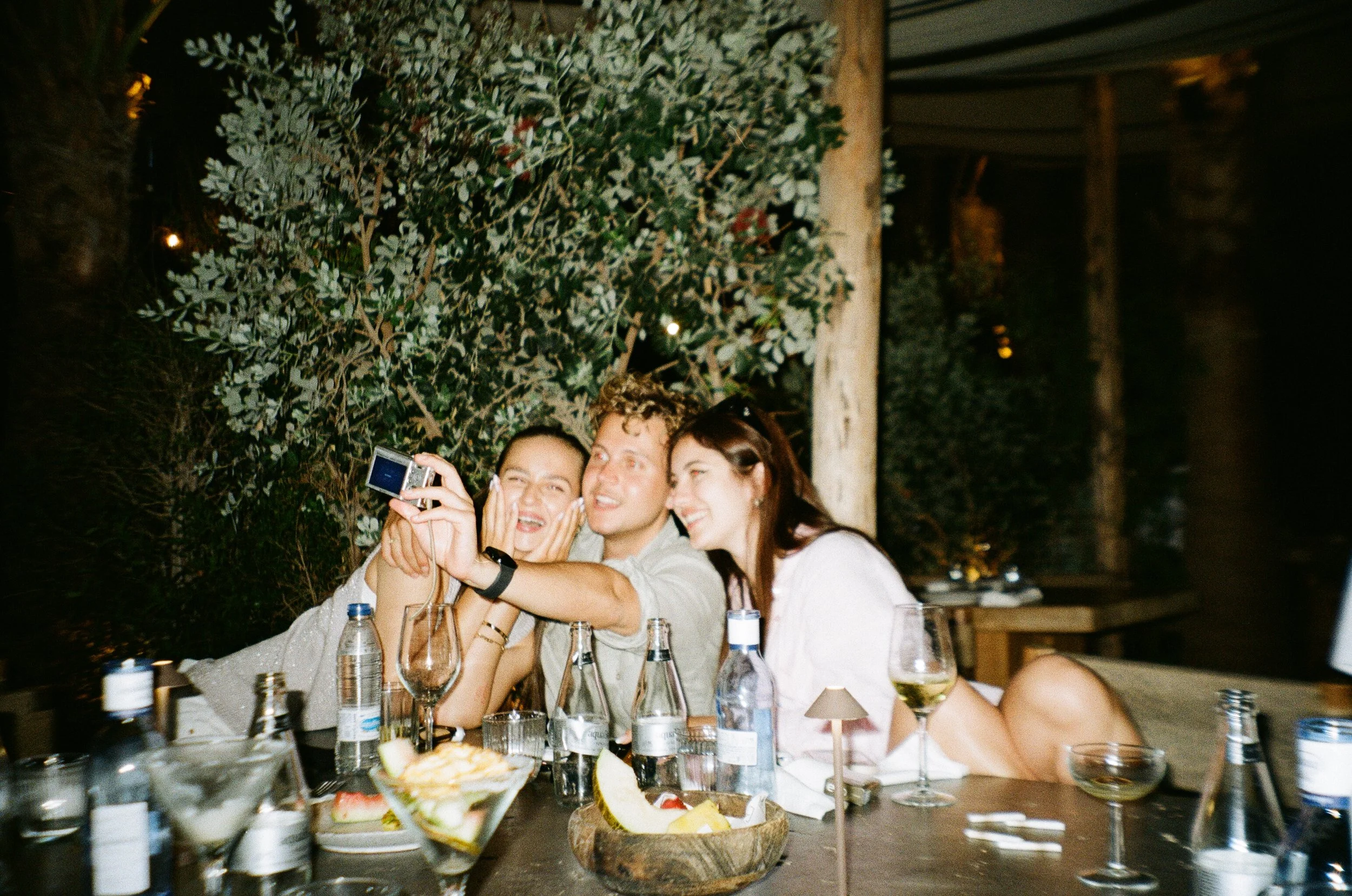 Group of people taking a selfie at a dinner table with various bottles and food, in an outdoor or garden setting at night.