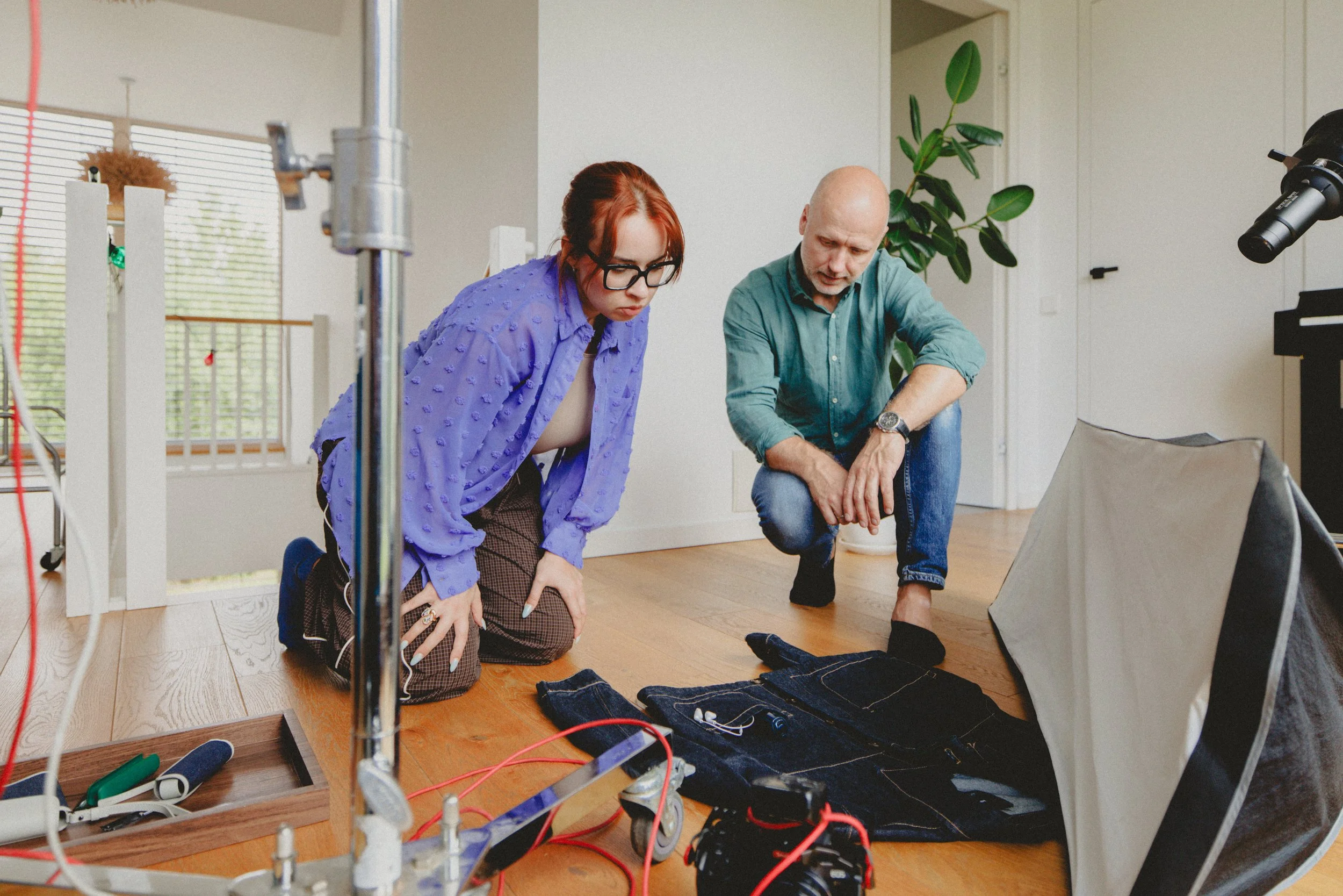 Two individuals, a woman with red hair and glasses, and a bald man, kneel on the wooden floor examining artwork or material on the ground in a well-lit room with large windows, a potted plant, and photographic equipment nearby.