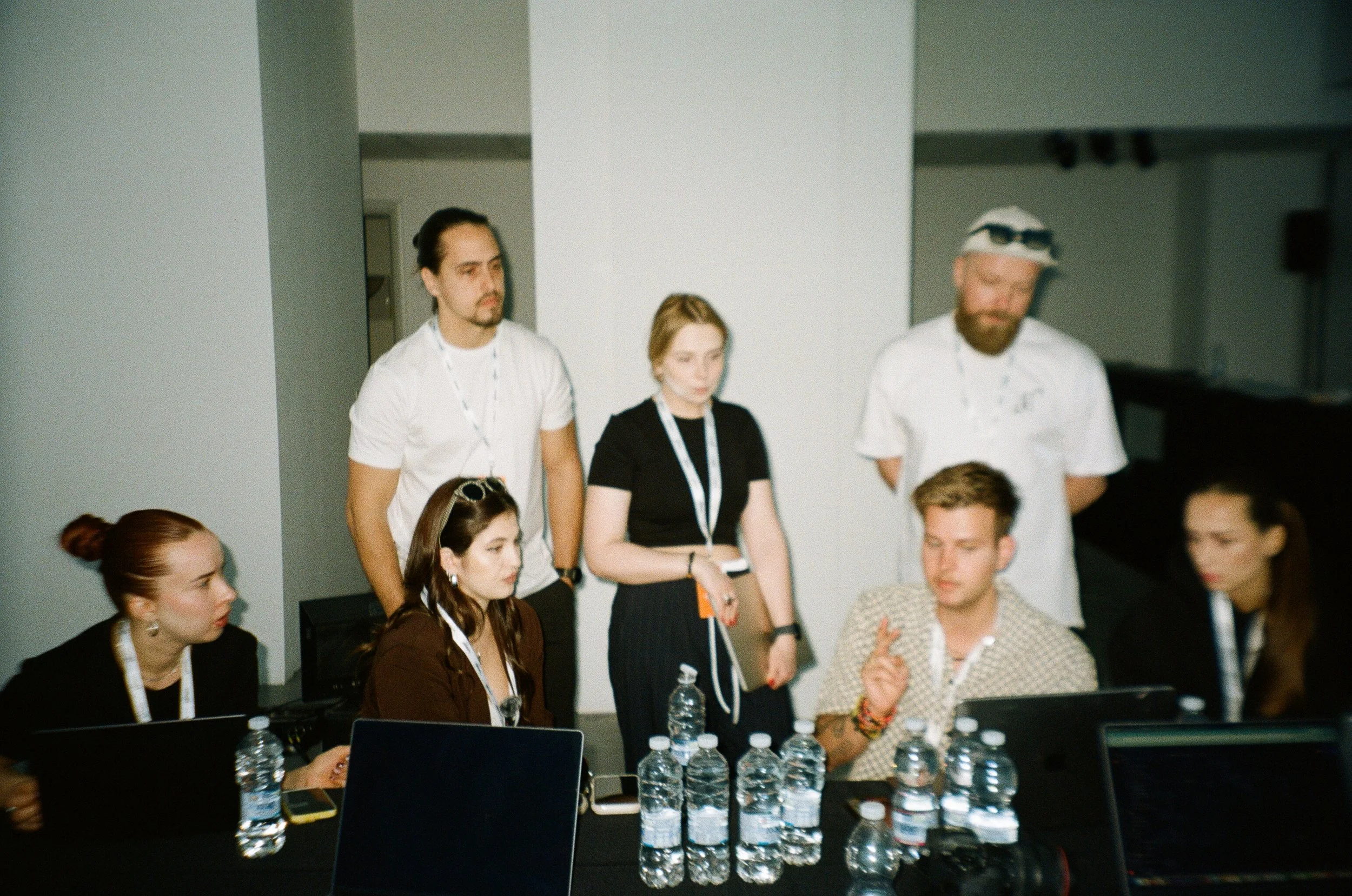 A group of people, some seated and some standing, gathered around a table with water bottles and laptops, in a room with white walls.