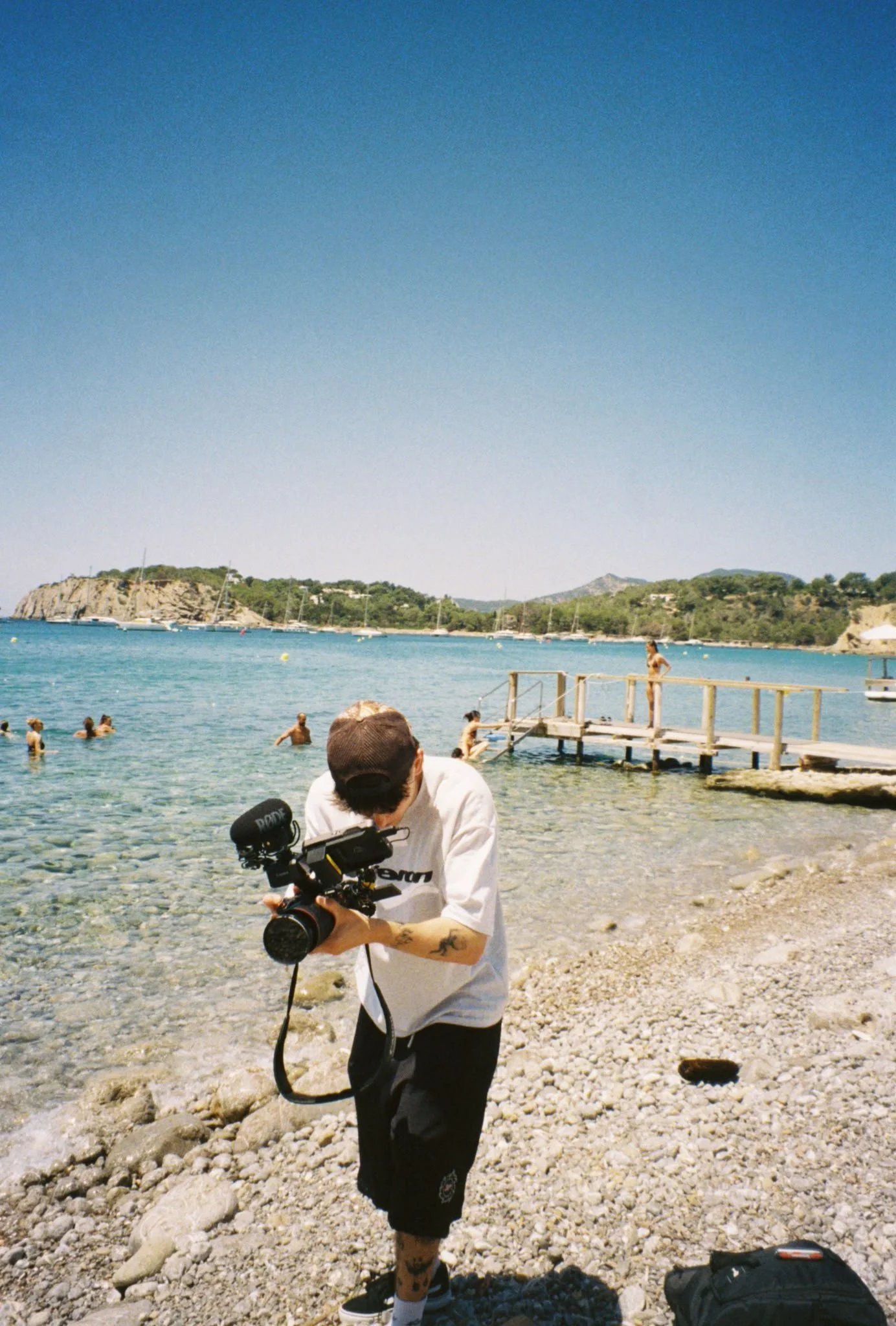 Person in white shirt and black shorts holding a camera near a rocky beach with people swimming, a wooden pier, boats, hills, and blue sky in the background.