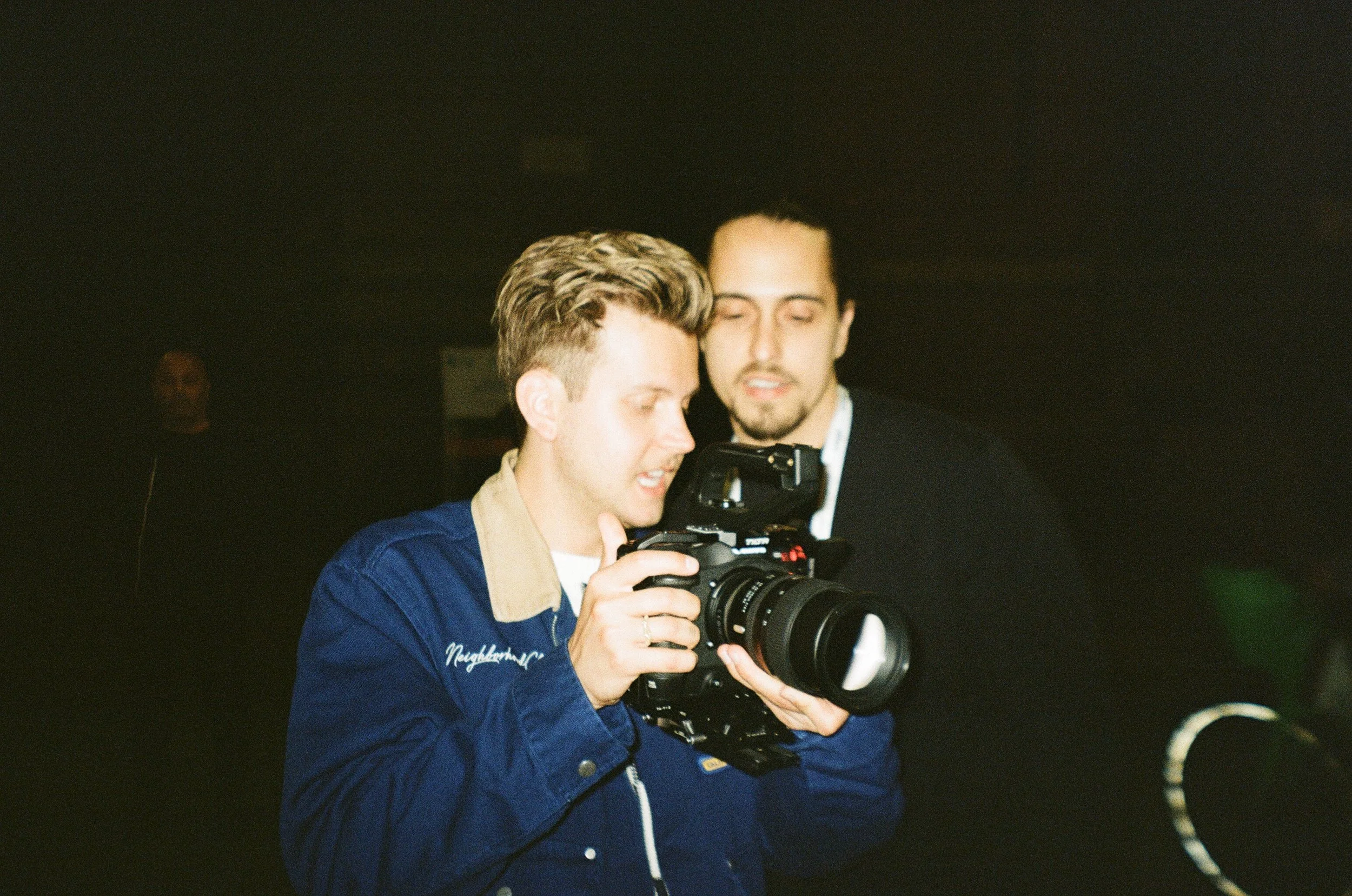 Two men looking at a camera, one holding it, in a dimly lit environment.