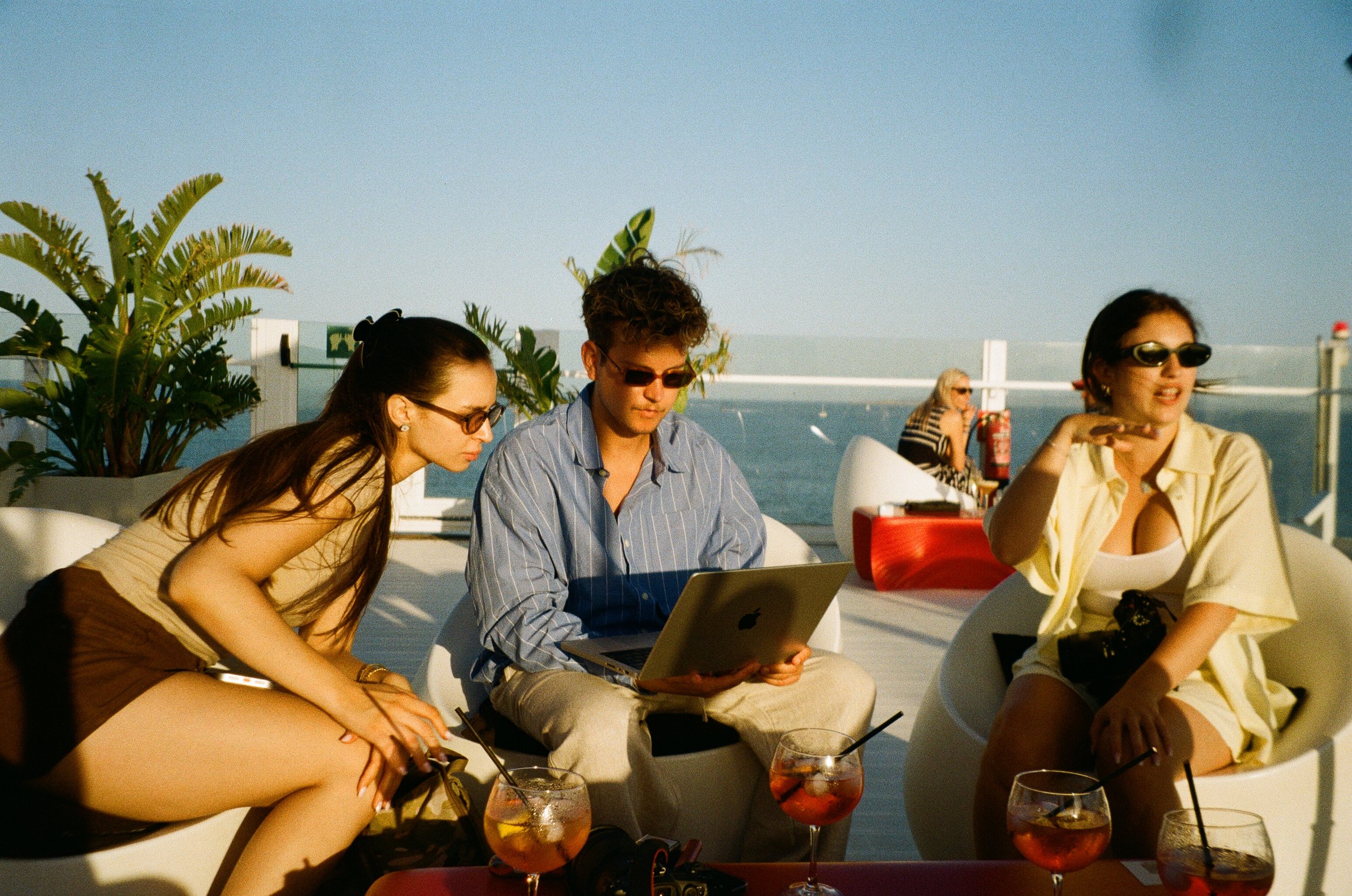 A group of young adults sitting outdoors at a rooftop venue, using laptops and enjoying drinks, with a view of the ocean and clear sky in the background.