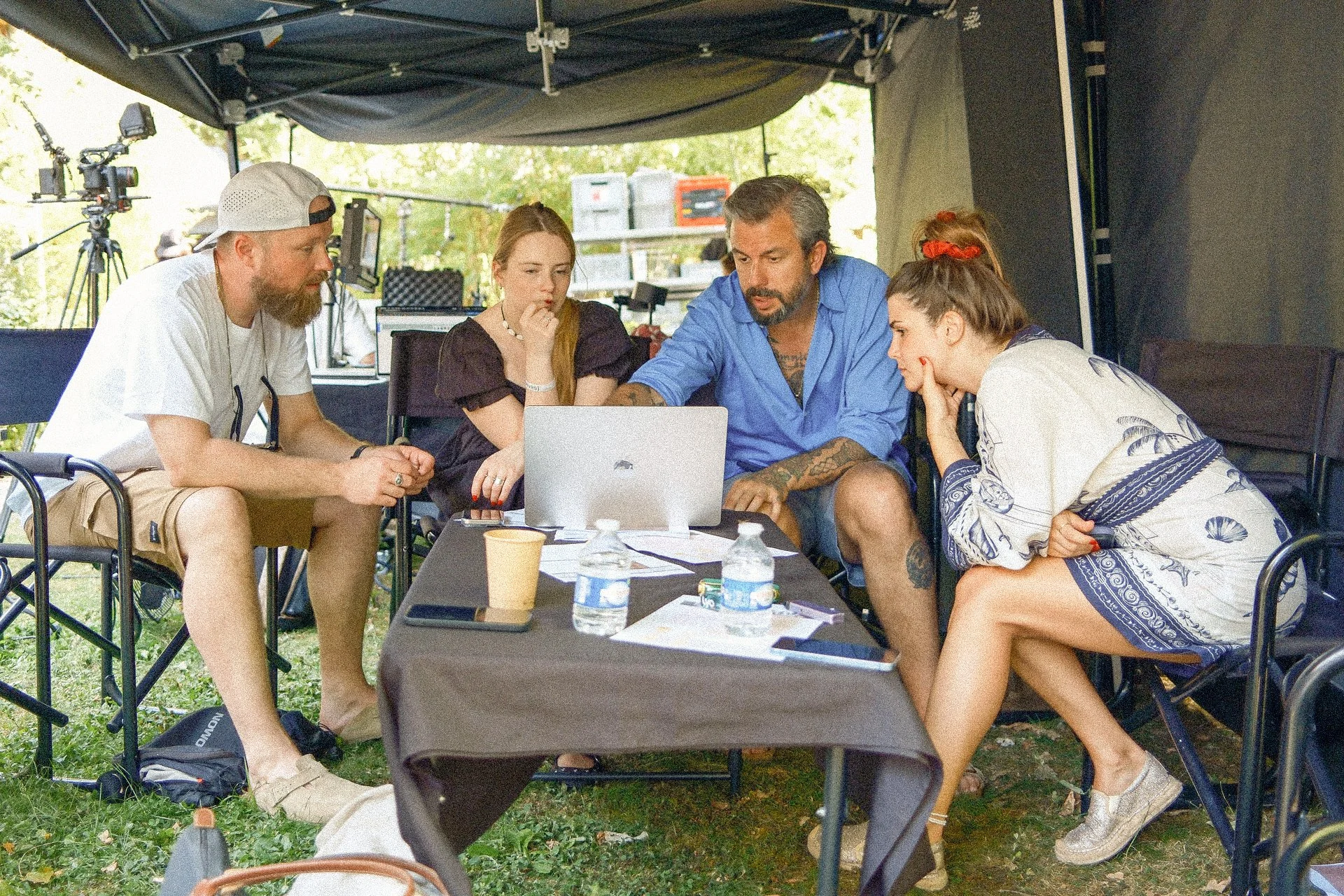 A group of five people sitting around a table under a canopy, looking at a laptop. There are water bottles, papers, and coffee cups on the table. The setting appears to be outdoors at a casual meeting or discussion.