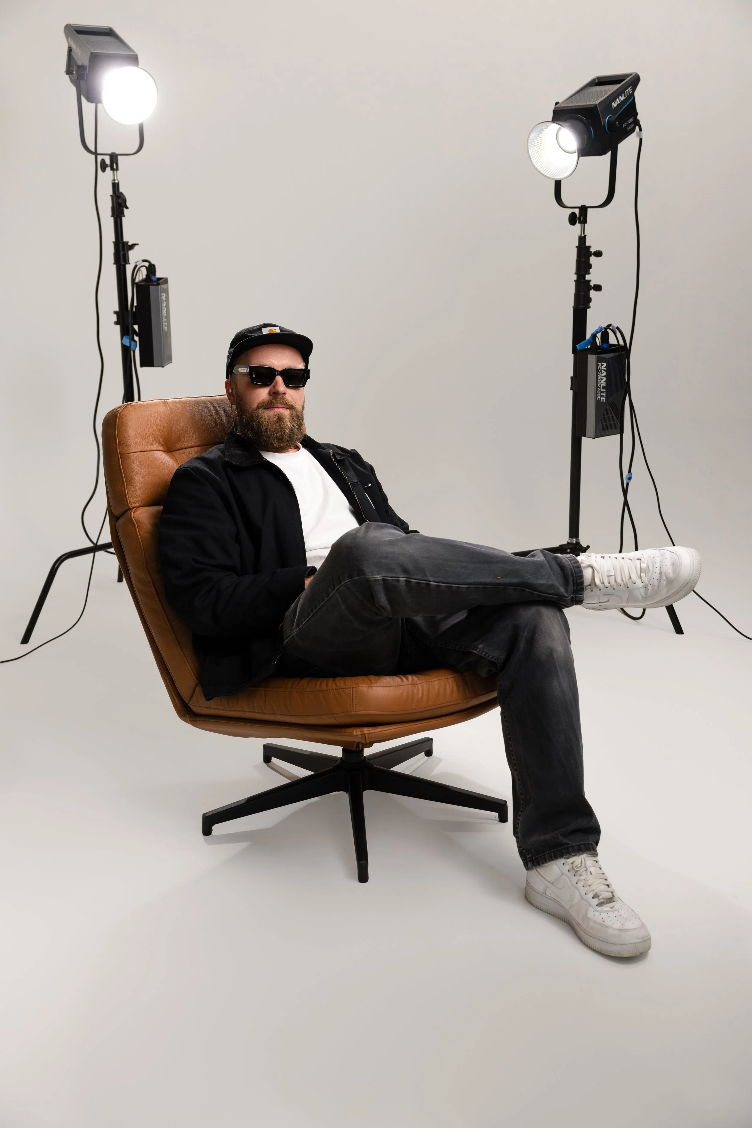A man with a beard, wearing sunglasses, a black baseball cap, a black jacket, and white sneakers, sitting in a tan office chair with his legs crossed, surrounded by studio lighting equipment on stands in a white studio setting.