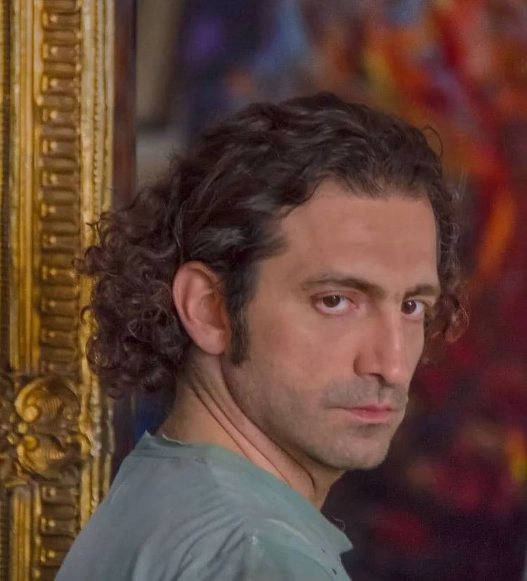 A man with curly dark hair, wearing a gray shirt, gazes over his shoulder with a serious expression, standing in front of a ornate gold-framed mirror with a colorful background.