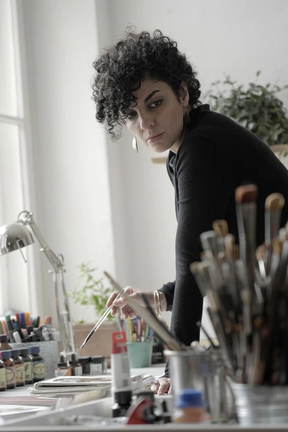 A woman with curly dark hair working at an art or craft workspace, holding a paintbrush and surrounded by art supplies, in a well-lit room with a potted plant in the background.