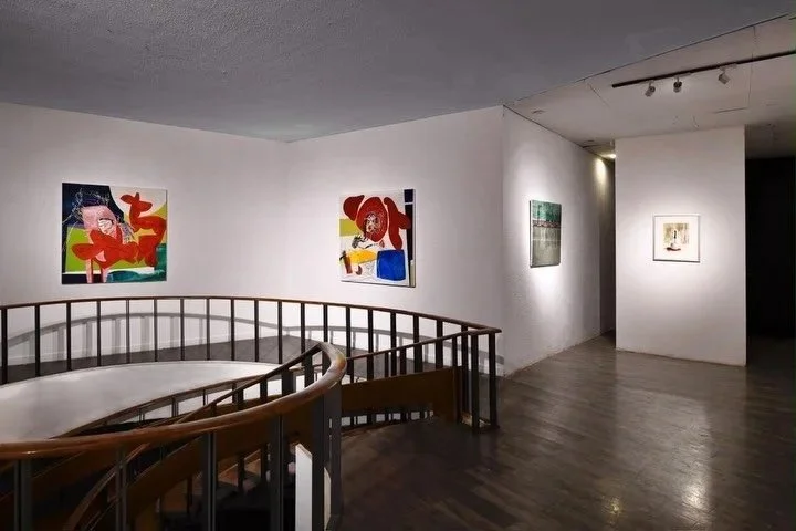 Art gallery with colorful abstract paintings on white walls and a curved wooden railing in the foreground.