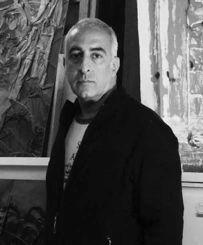 A man with short gray hair and a serious expression, wearing a dark jacket and white shirt, standing indoors near an abstract painting on the wall.