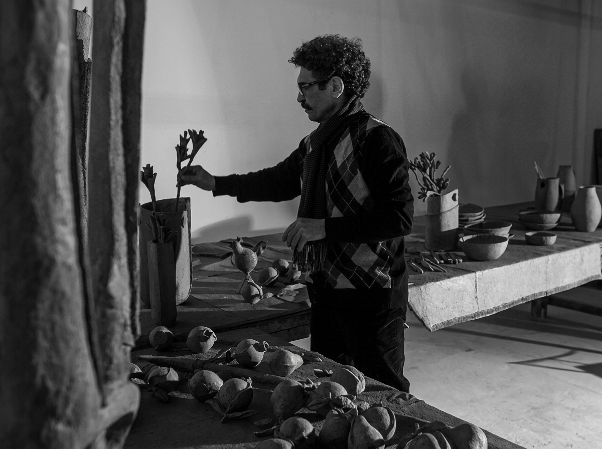 A man with curly hair and glasses arranging flowers in a vase in an art studio with pottery and sculptures on the table.