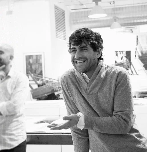 A smiling man in a light-colored sweater talking in an indoor setting with other people and artwork in the background.