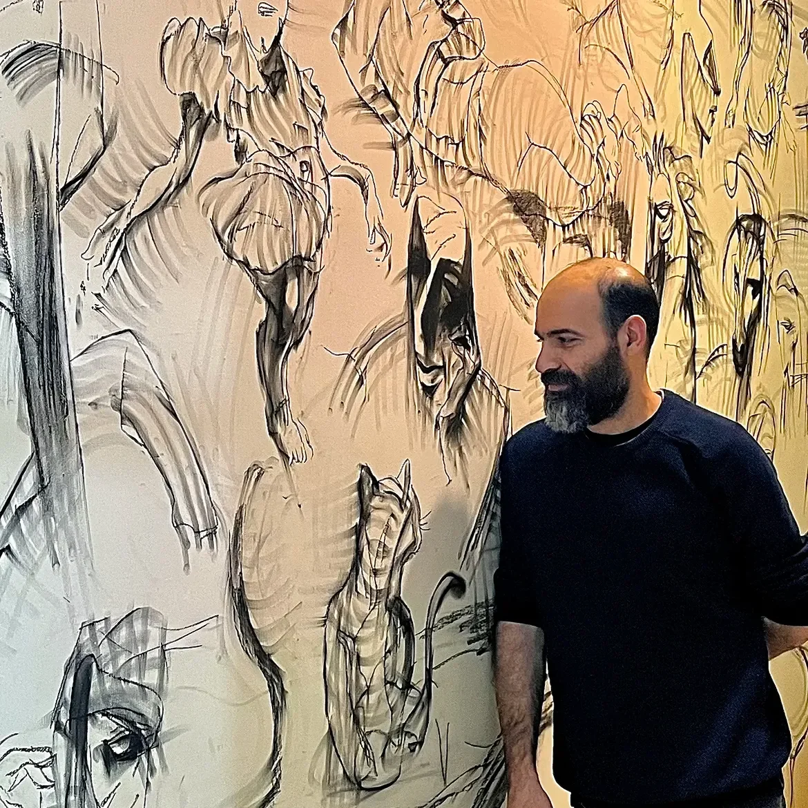 Man with a beard standing in front of a wall with a large sketch of a horse and other animals.
