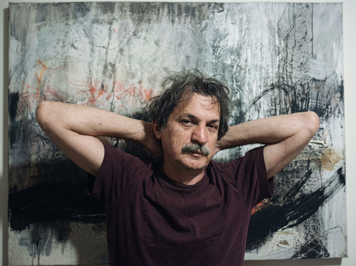 A man with gray, wavy hair and a mustache stands with arms behind his head in front of an abstract painting with dark, white, and some red strokes.