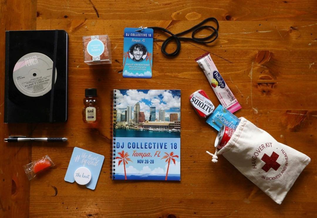 Collection of items on a wooden surface, including a vinyl record, a marker, a notebook with cityscape cover, a pass badge, a small bottle of medication, a small container of dental floss, a lanyard, a ginger chew, gel packets, canned beverages in a tote bag with a red cross, and some small packaged items.