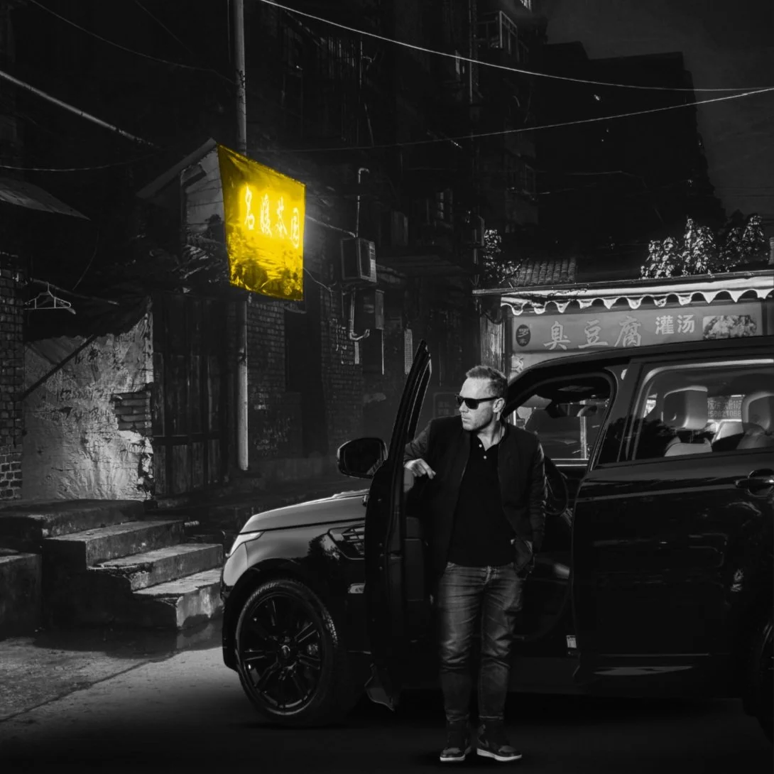 A man wearing sunglasses and a black jacket stands next to a black SUV on a city street at night, with Asian signage and illuminated yellow sign in the background, in black and white.