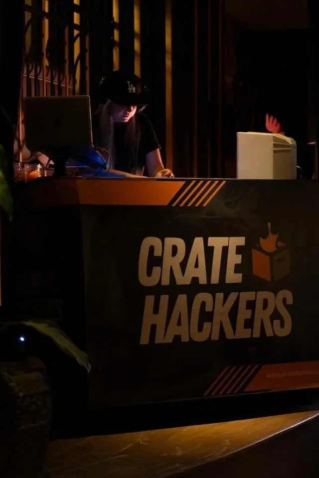 A person working at a computer behind a counter with a sign that reads 'CRATE HACKERS' at a dark, indoor event.