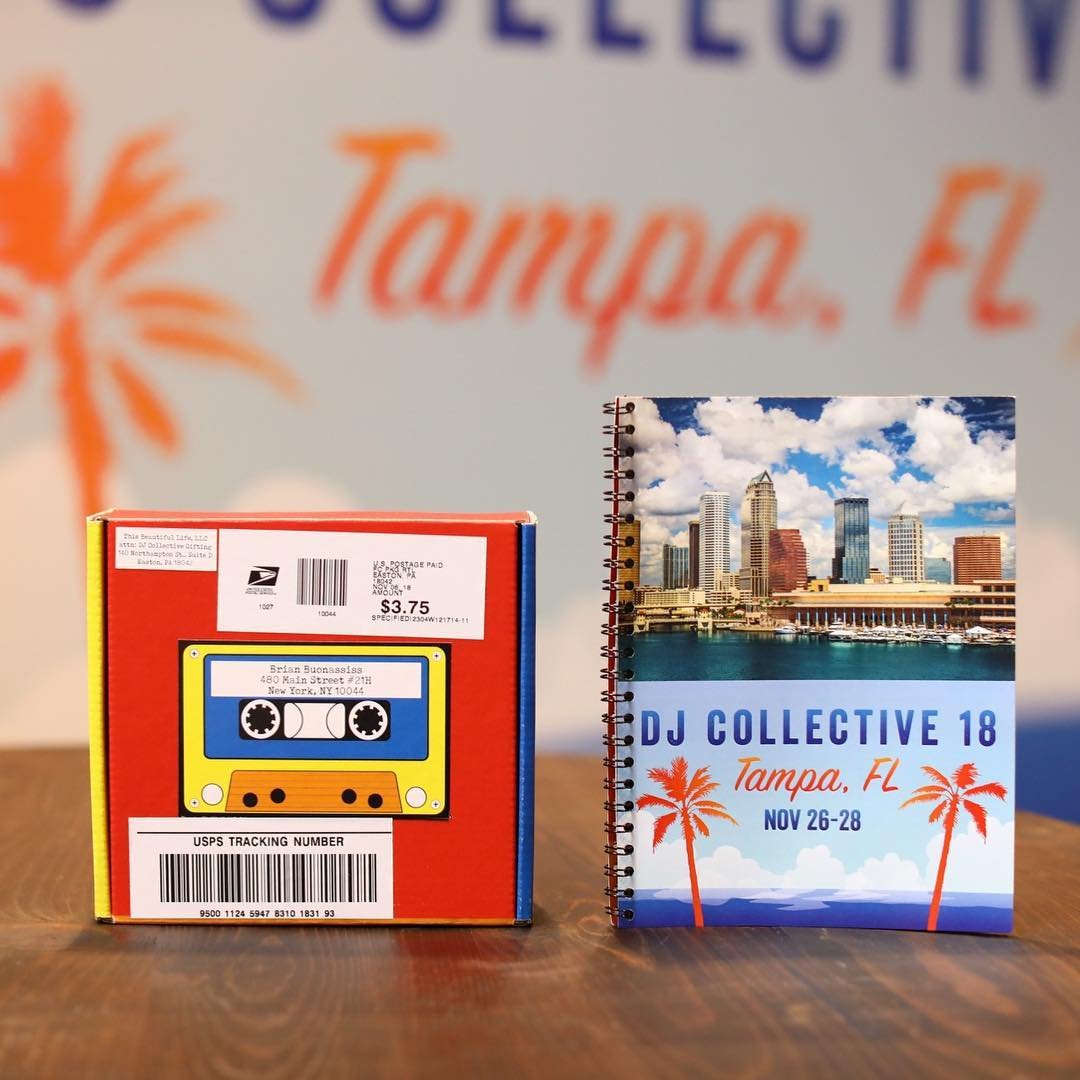 A postage box and a spiral notebook on a wooden table. The box features a colorful cassette tape illustration and a mailing address sticker. The notebook has a cover photo of Tampa's skyline with clouds and palm trees, and text about DJ Collective 18 event in Tampa, FL from November 26-28.