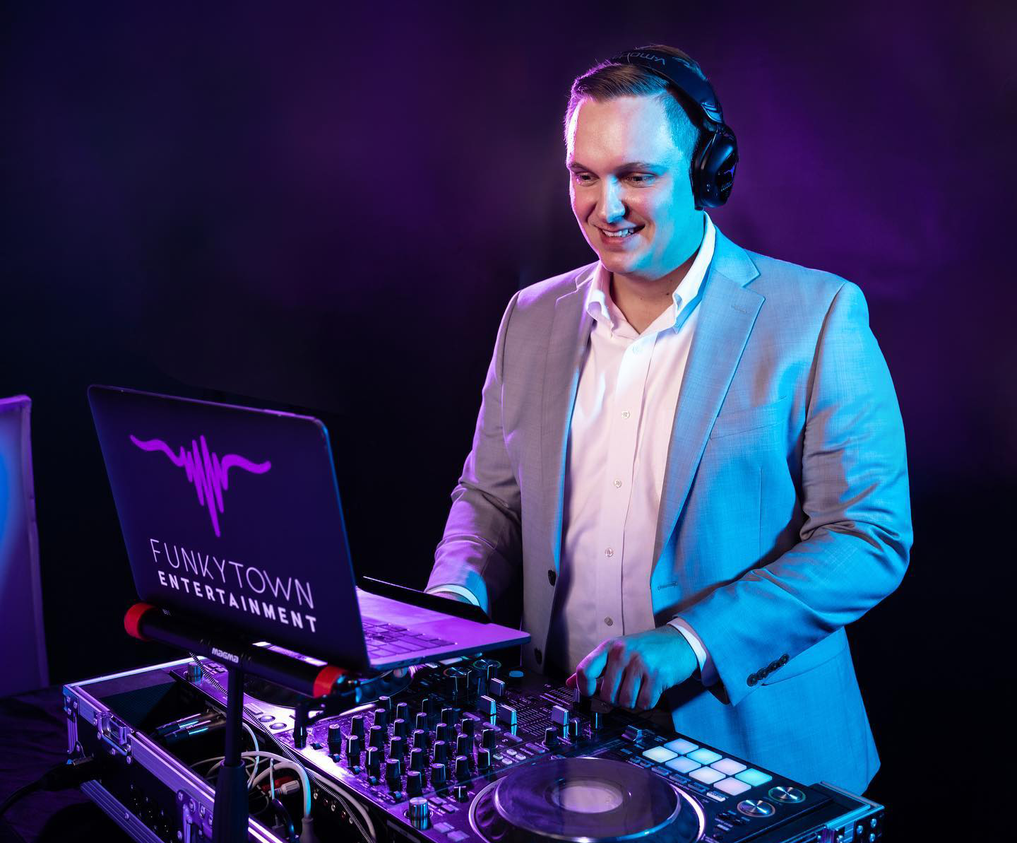 A man in a light gray suit and white shirt is DJing with headphones on in a dark, colorful setting. His DJ equipment is set up with a laptop that has the logo and text 'FUNKYTOWN ENTERTAINMENT' on it.