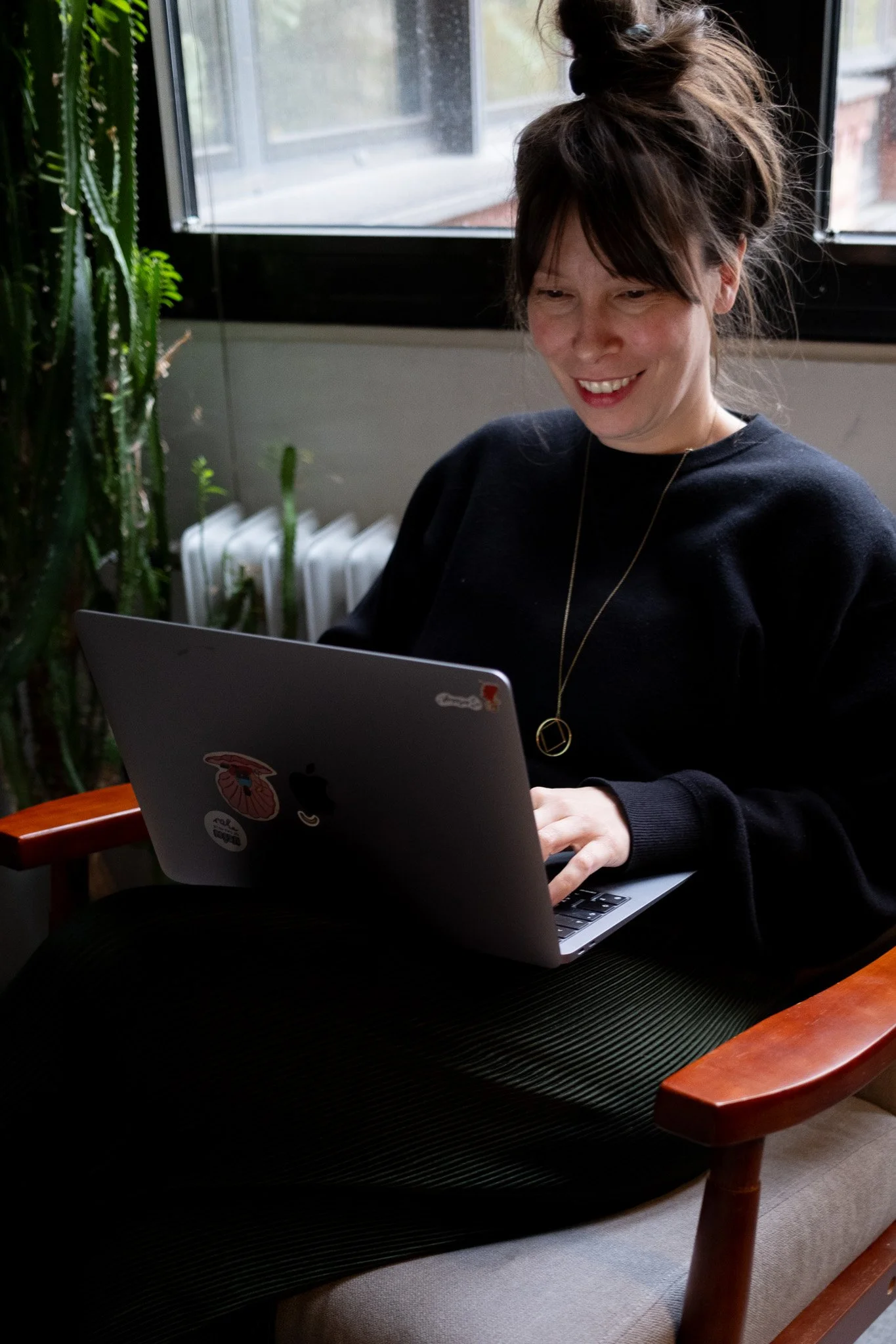 Eine lächelnde Frau sitzt an einem Tisch und arbeitet an einem Laptop, während sie in einen Bildschirm schaut